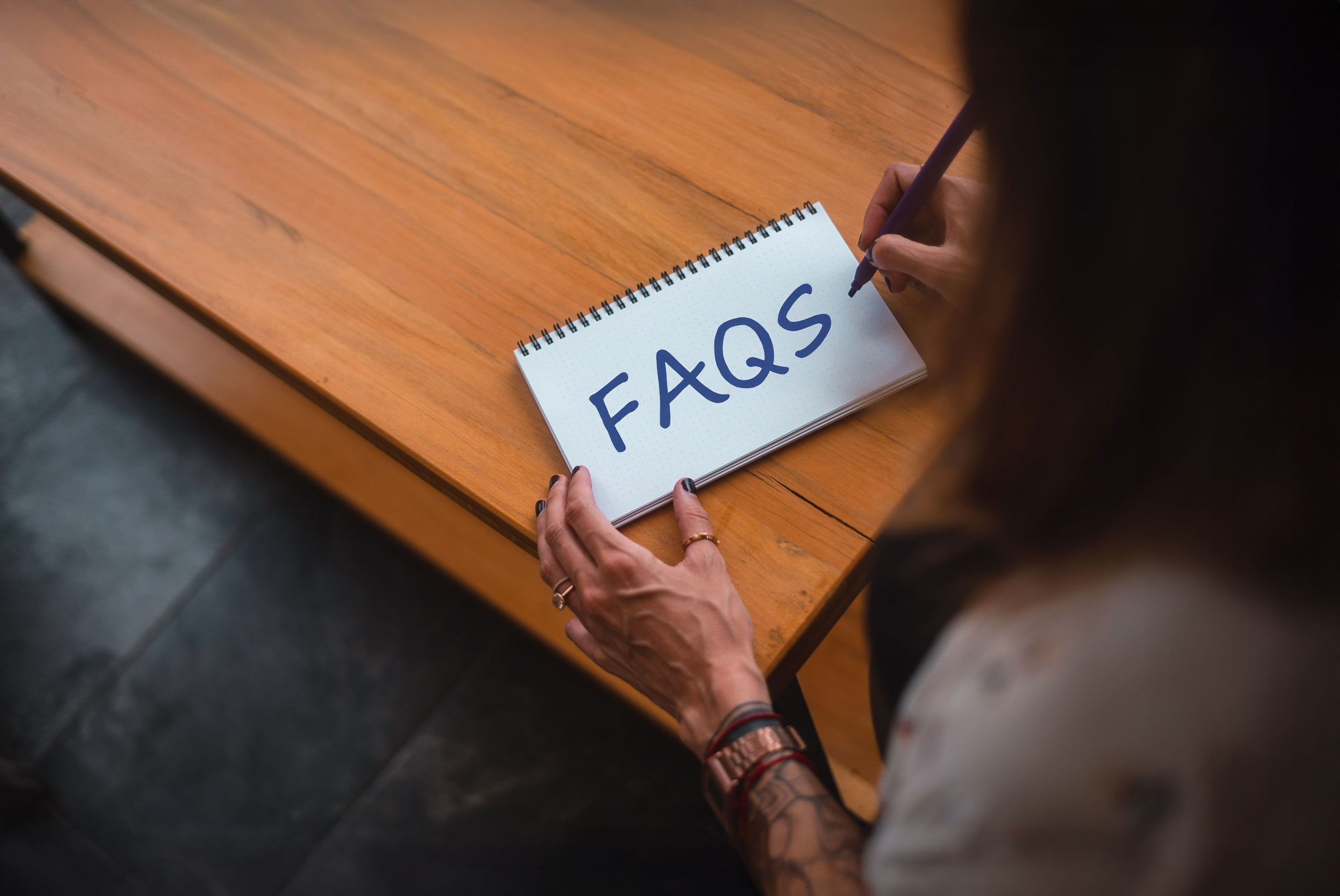 A person writes "FAQS" in blue on a spiral notepad at a wooden desk. 