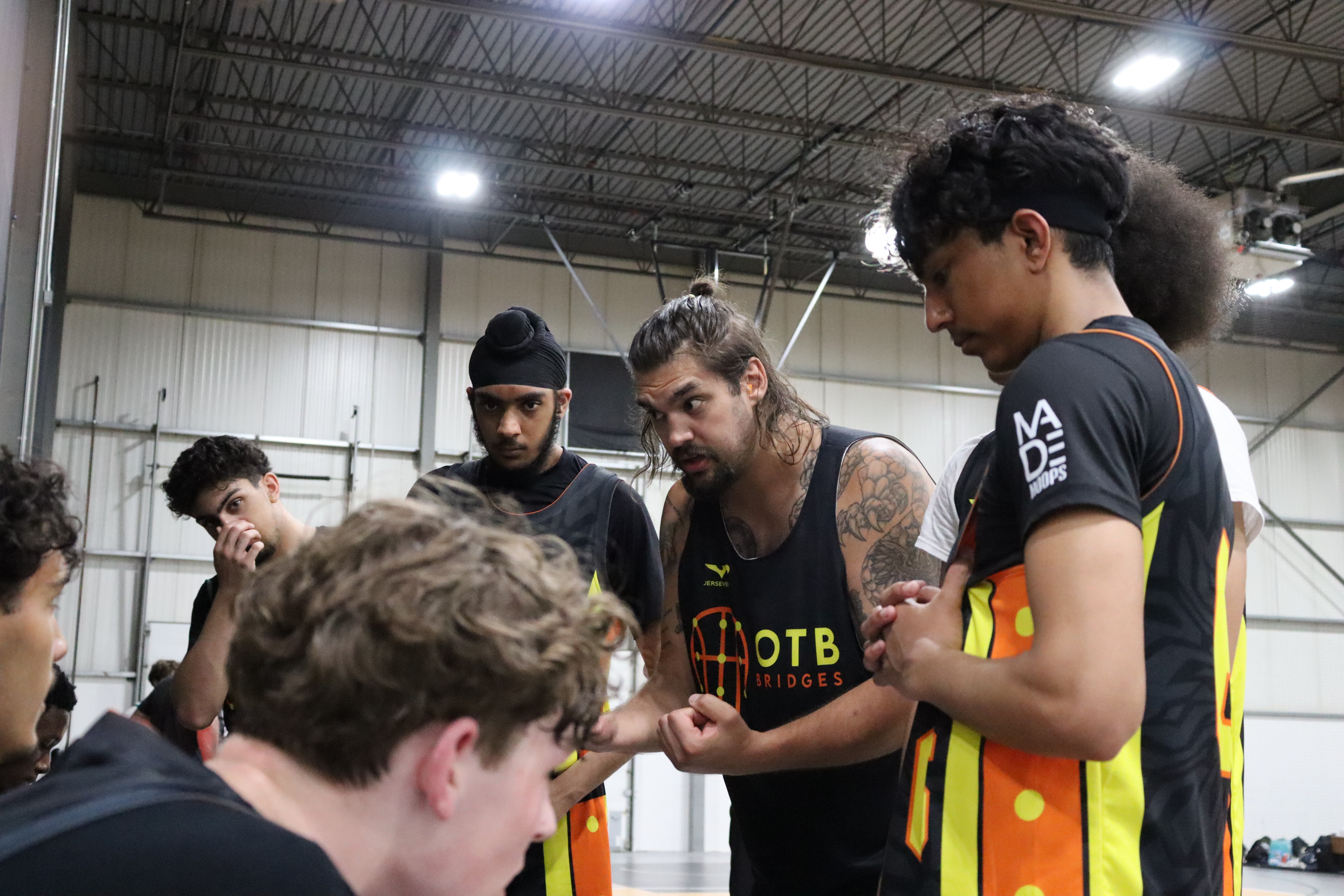 A youth basketball player in an orange jersey dribbling aggressively past a defender during a game, with a referee and spectators visible in the background on an indoor court.