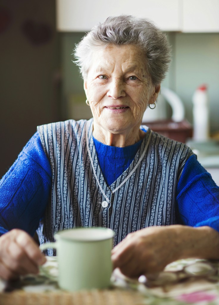 A cheerful elderly woman sitting at a table with a cup in her hands, wearing a blue sweater and a striped vest, exuding warmth and contentment in a cozy indoor setting.