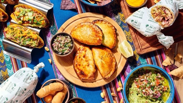 This vibrant image showcases a colorful spread of Mexican cuisine, featuring a wooden platter of golden-brown empanadas, a bowl of fresh guacamole topped with diced vegetables, tacos filled with seasoned meat and greens, burritos wrapped in branded paper, and a small dish of salsa, all set against a festive woven tablecloth.