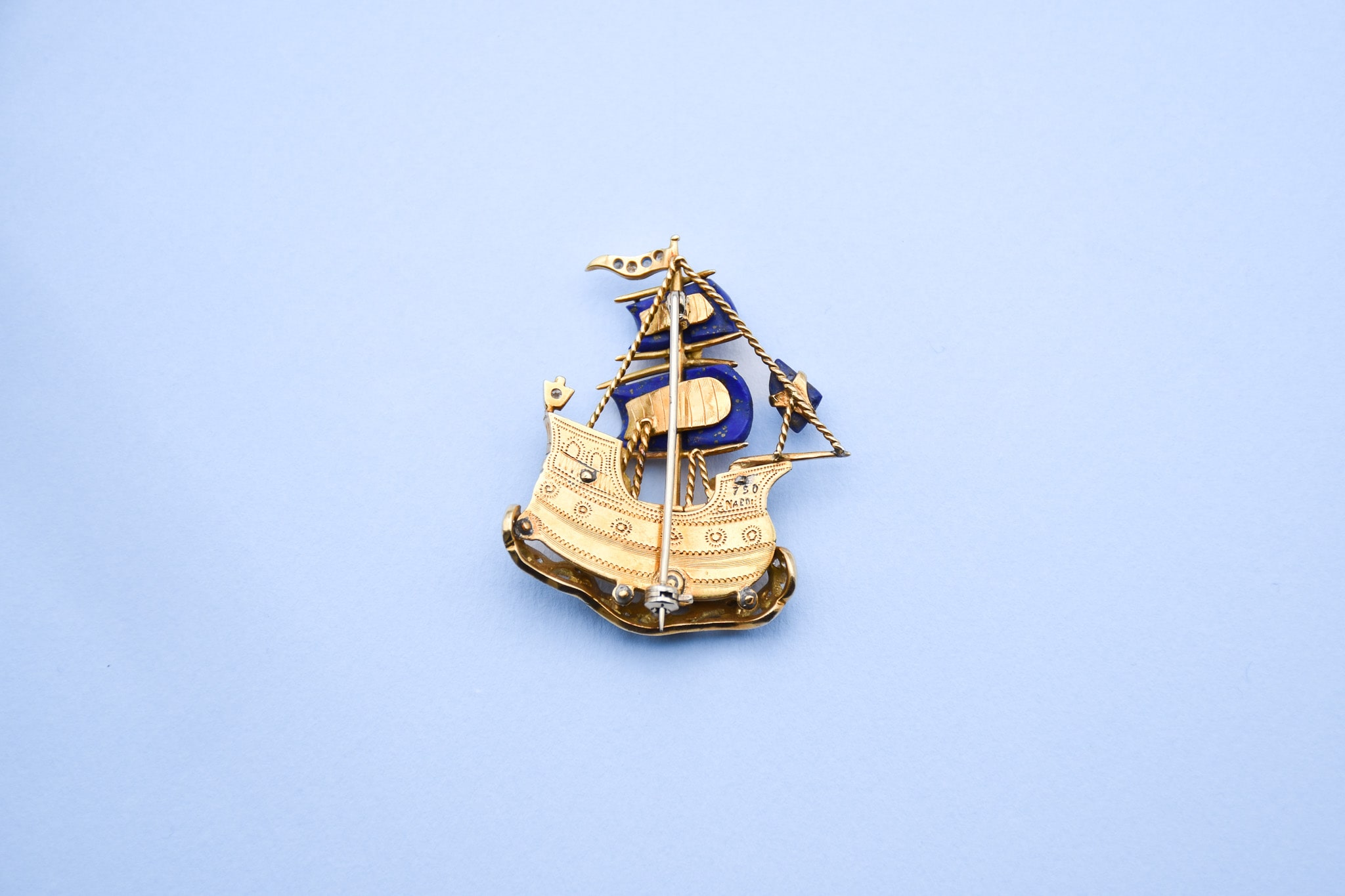 Vue de dos d'une broche représentant un galion naviguant sur les eaux dont les voiles sont en lapis-lazuli et la coque en émail blanc ornée de petits diamants