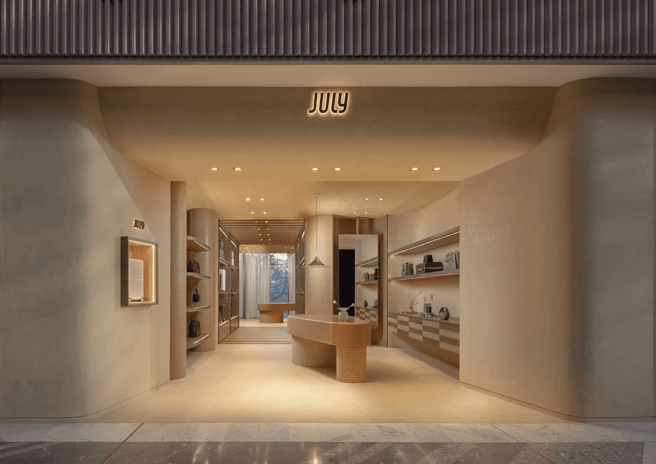 Minimalist retail storefront with illuminated “JULY” sign above the entrance, featuring a warm-toned interior with wooden display tables, shelving, and soft lighting.