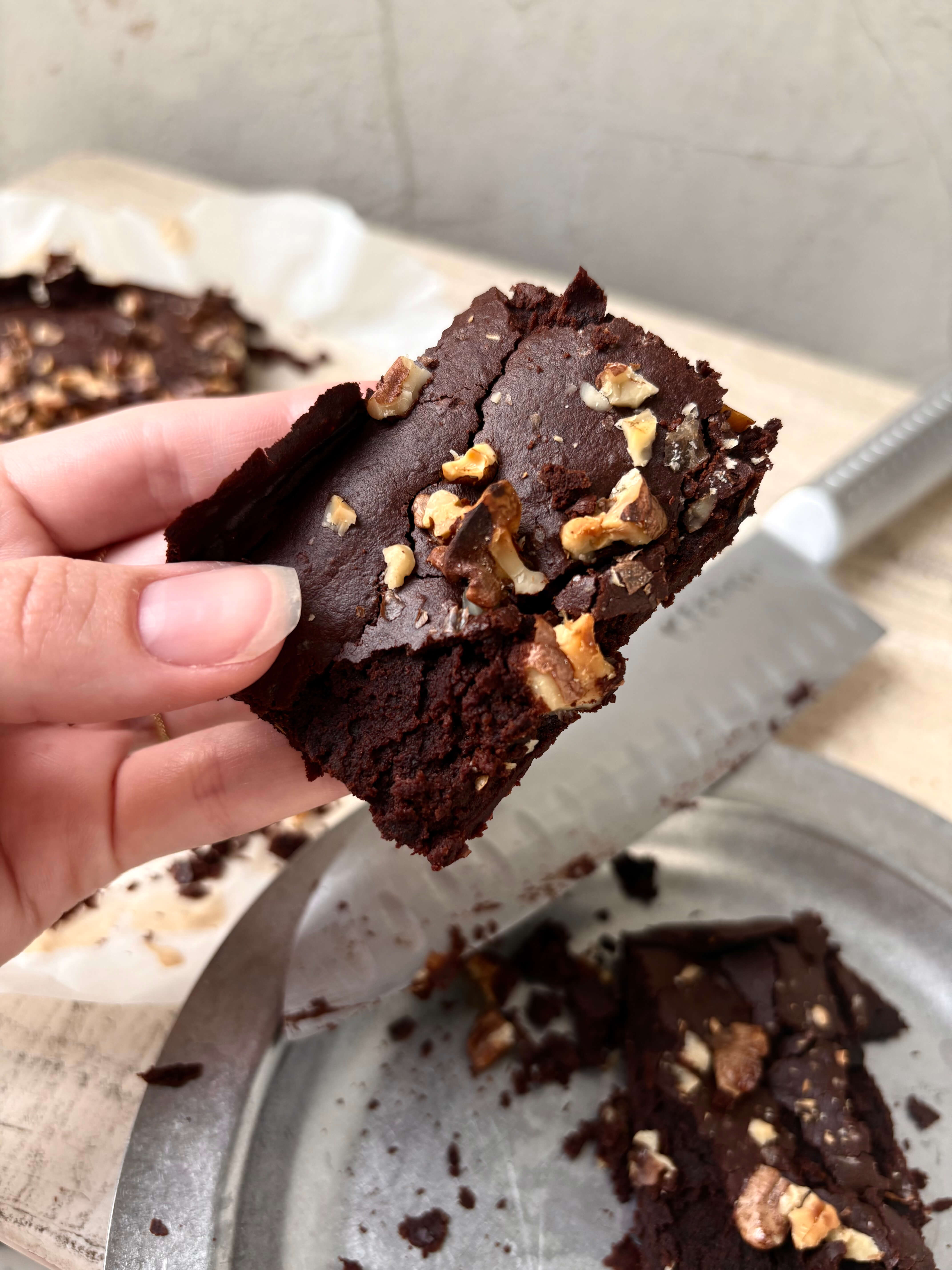 Close-up of a rich, fudgy kidney bean brownie topped with chopped walnuts, held by hand above a tray of sliced brownies. A nutrient-dense chocolate snack with added fibre, iron, protein, and healthy fats.