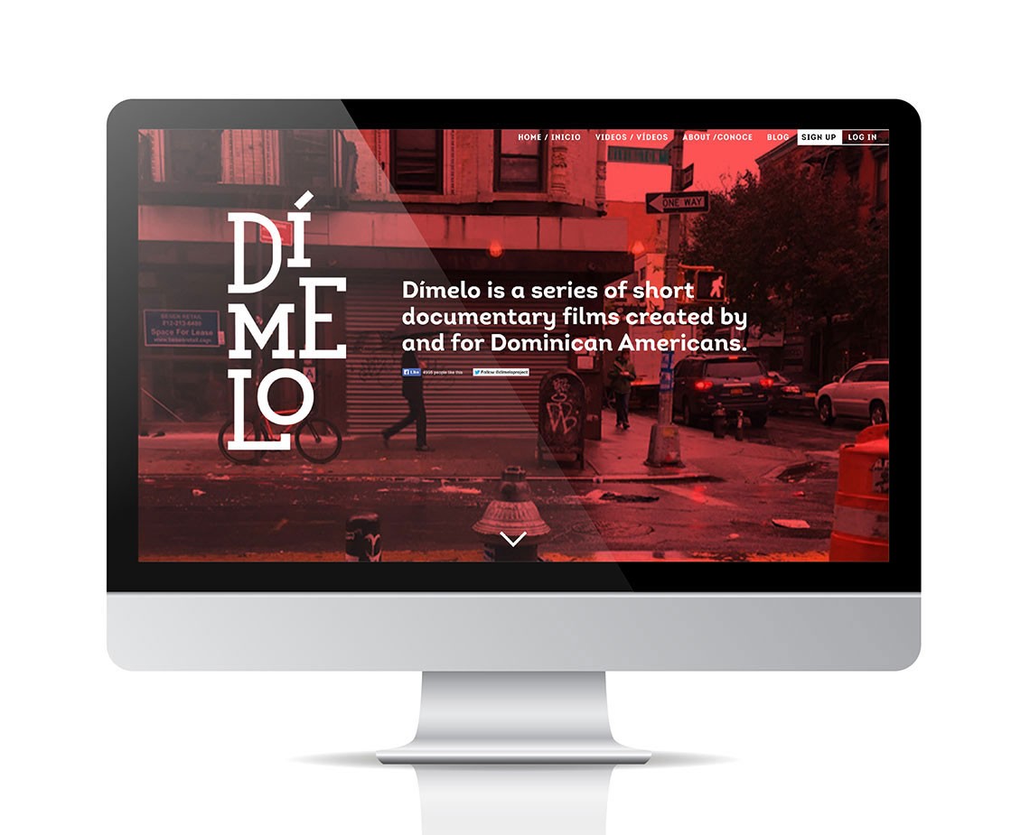 A computer monitor displays the website for Díme Lo, a series of short documentary films created for Dominican Americans. The website features a red-toned background with text and graphics, including the title “Díme Lo” in large white letters. The navigation bar includes links for “Home,” “Inicio,” “Vídeos,” “About/Conoce,” “Blog,” and “Sign Up,” along with a “One Way” button. The monitor is positioned on a white surface against a white background.