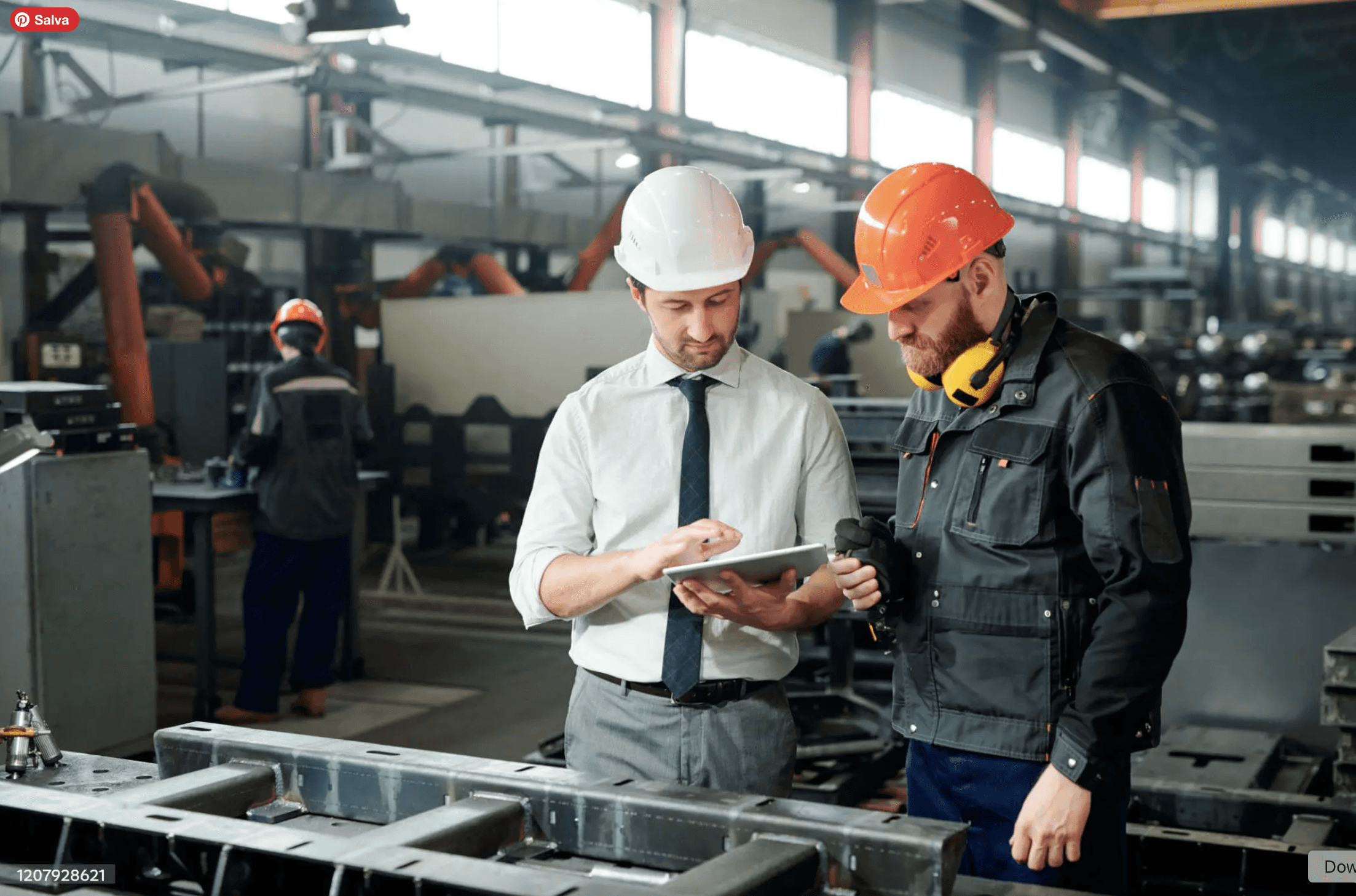 Dettaglio professionale di un ingegnere in camicia e cravatta con elmetto bianco che collabora con un operaio specializzato in tuta da lavoro ed elmetto arancione, mentre consultano insieme un tablet all'interno di un capannone industriale.
