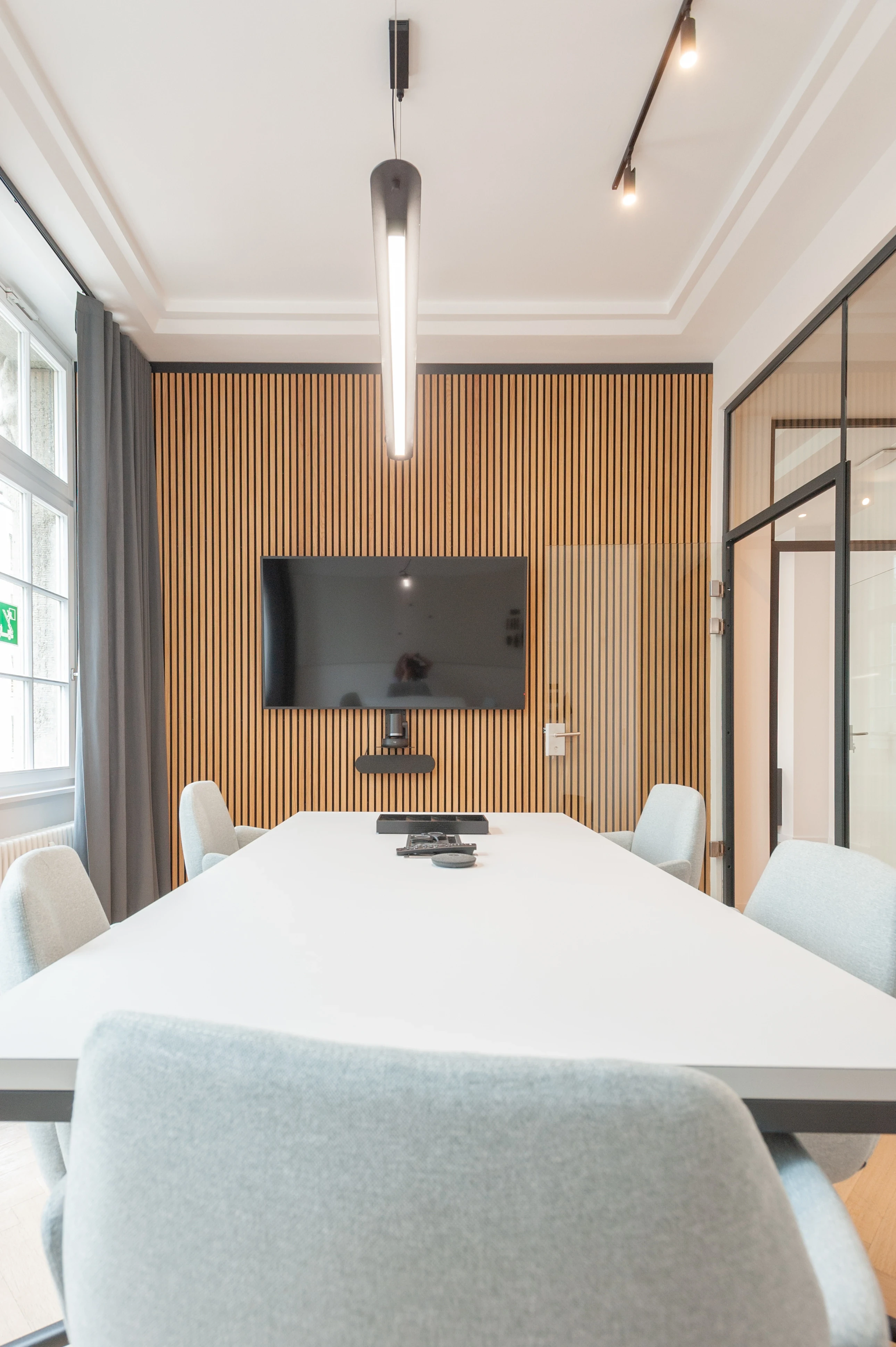 Video conference room at AI START-UP BERLIN with a white table, light blue upholstered chairs, a TV on an oak slat wall, a vertical black pendant light and stucco ceiling mouldings.