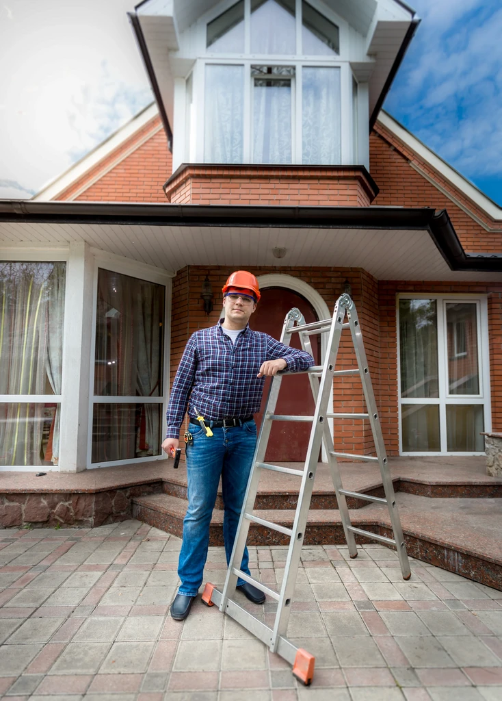 A construction worker in a hard hat stands confidently beside a ladder in front of a two-story brick house with large windows on a bright day.