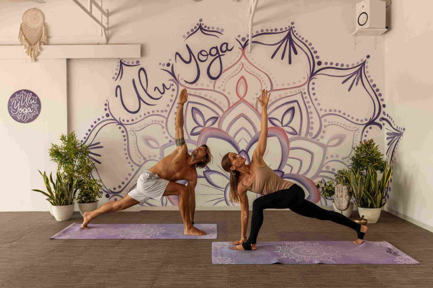 Male and female students practicing partner yoga flow in front of the hand-painted mandala wall at Ulu Yoga studio Bali.