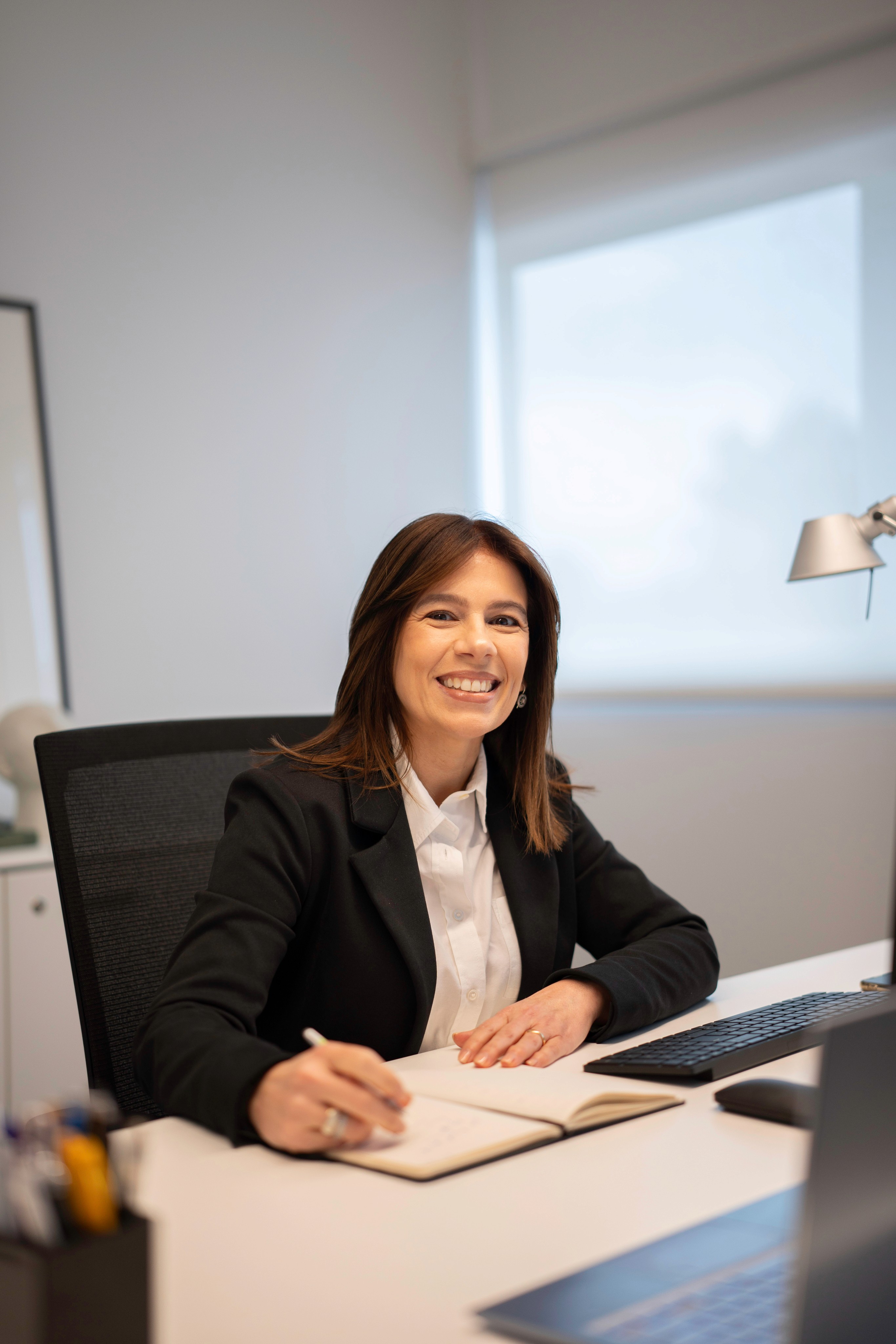 Mulher empresária sorrindo em um ambiente de escritório moderno, vestida com blazer preto e camisa branca, transmitindo confiança e profissionalismo.