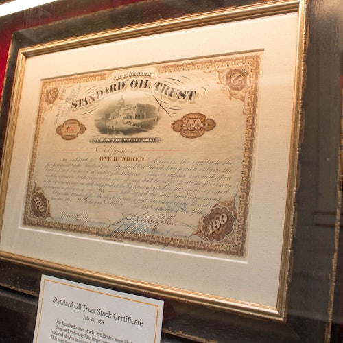 A framed Standard Oil Trust stock certificate displayed on a wall with a descriptive plaque beneath it.