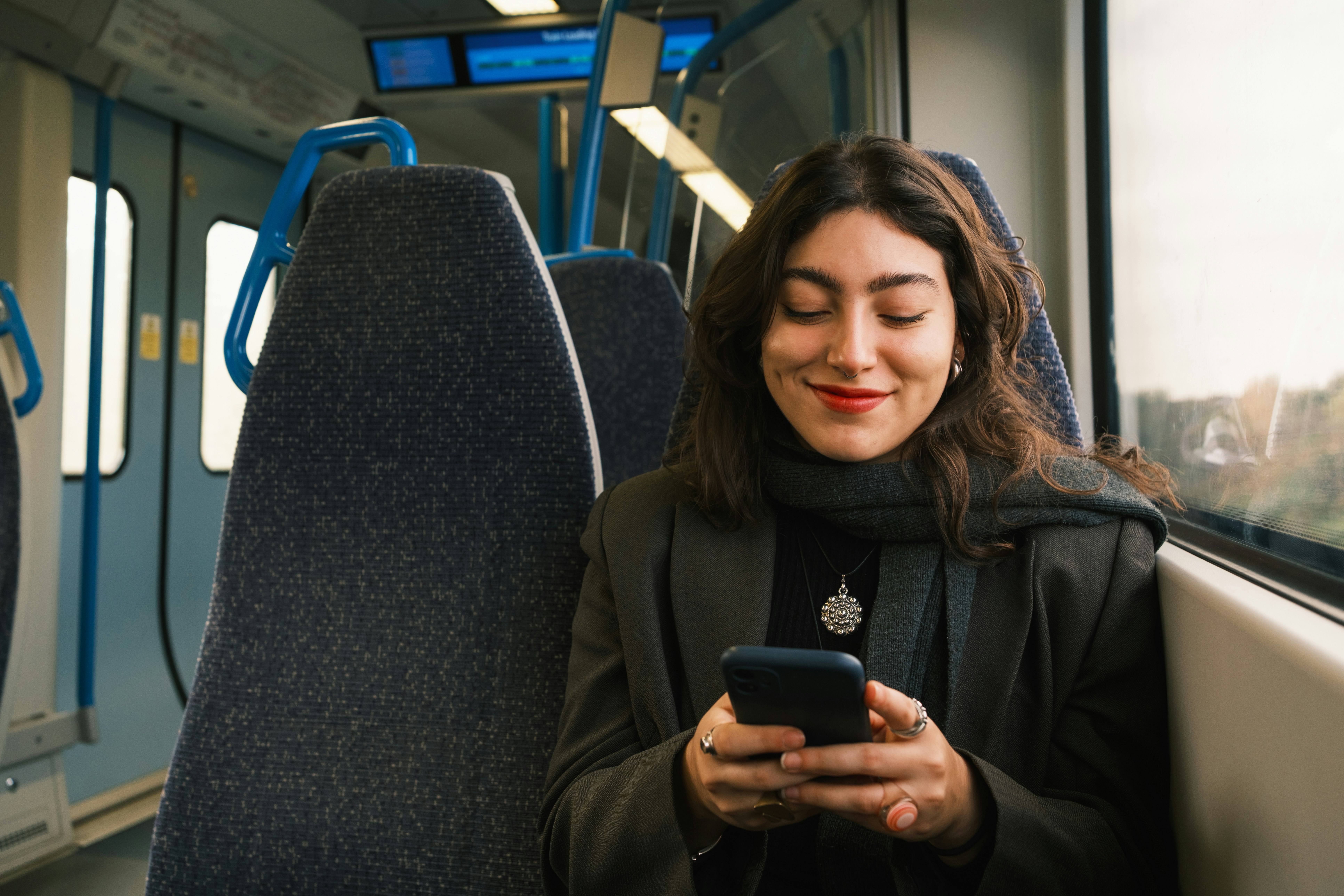 Mobiles Trade-In Portal für Kunden: Eine Frau nutzt ihr Smartphone in einer Straßenbahn, um über das digitale Kundenportal einen Ankauf zu tätigen. Das Display zeigt intuitive Bedienelemente, während im Hintergrund eine europäische Stadtkulisse durch das Fenster erkennbar ist.