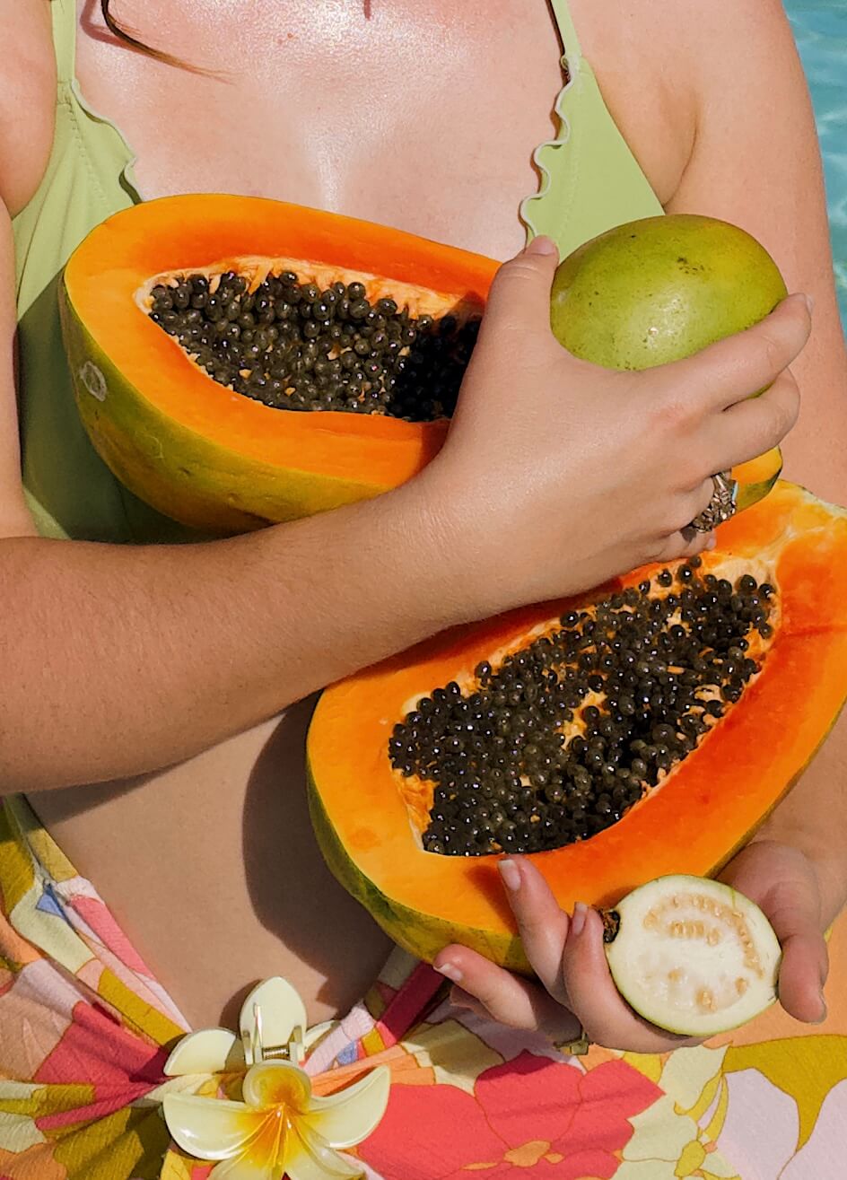 a woman in a chartreuse bikini top and pink and yellow sarong holding a large papaya cut in half, a mango, and half a guava