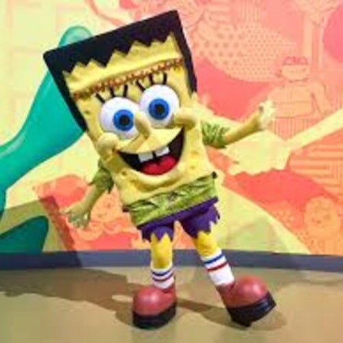 一個打扮成黃色卡通角色的人，擁有大藍眼睛、紅色鞋子和綠色襯衫，正站在一面多彩的牆前。