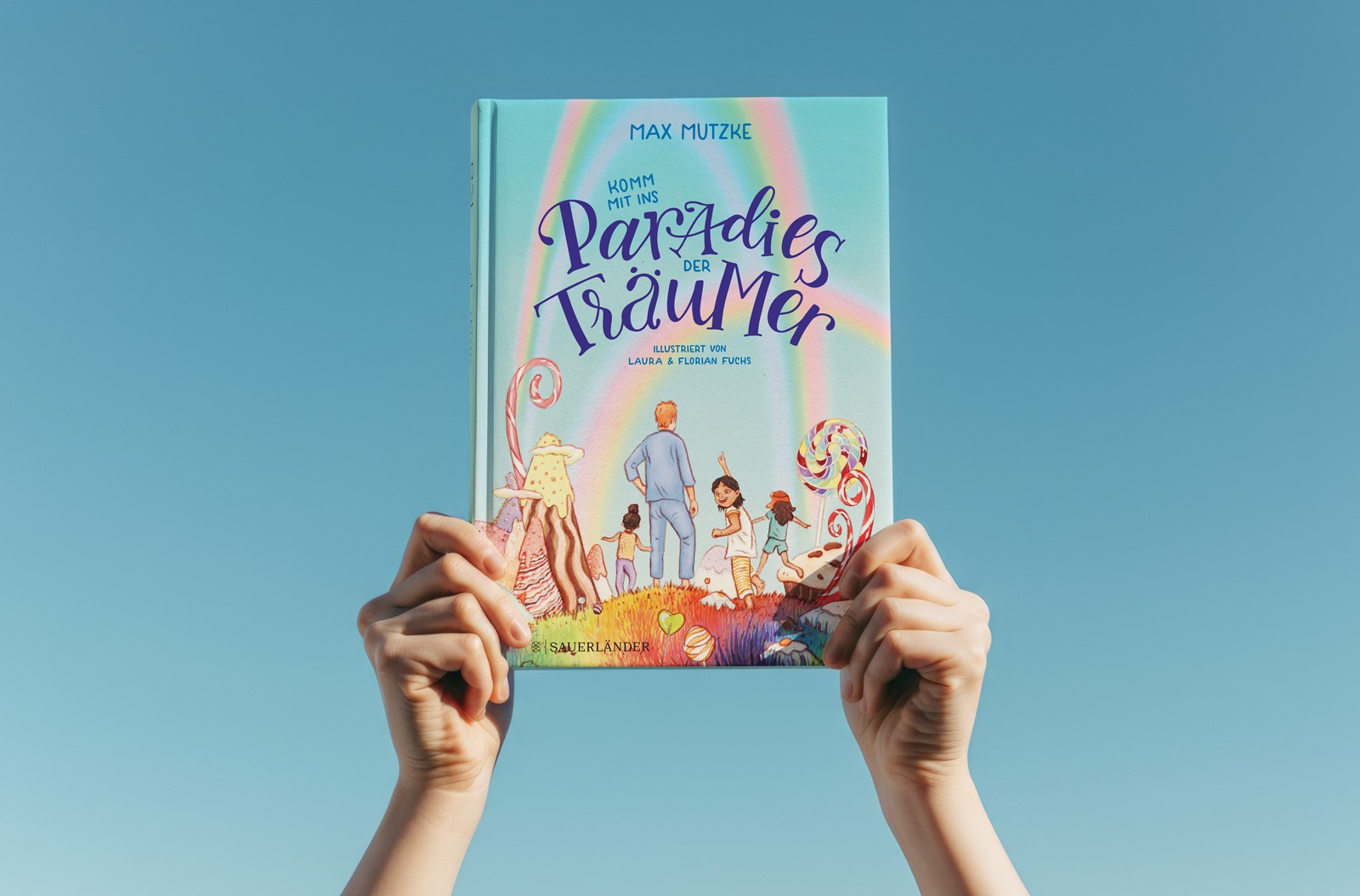 Hände halten das Kinderbuch „Paradies der Träumer“ in die Luft. Das Cover zeigt handgestaltetes Lettering des Titels, verspielte Figuren und einen Regenbogen, der eine Traumwelt darstellt.