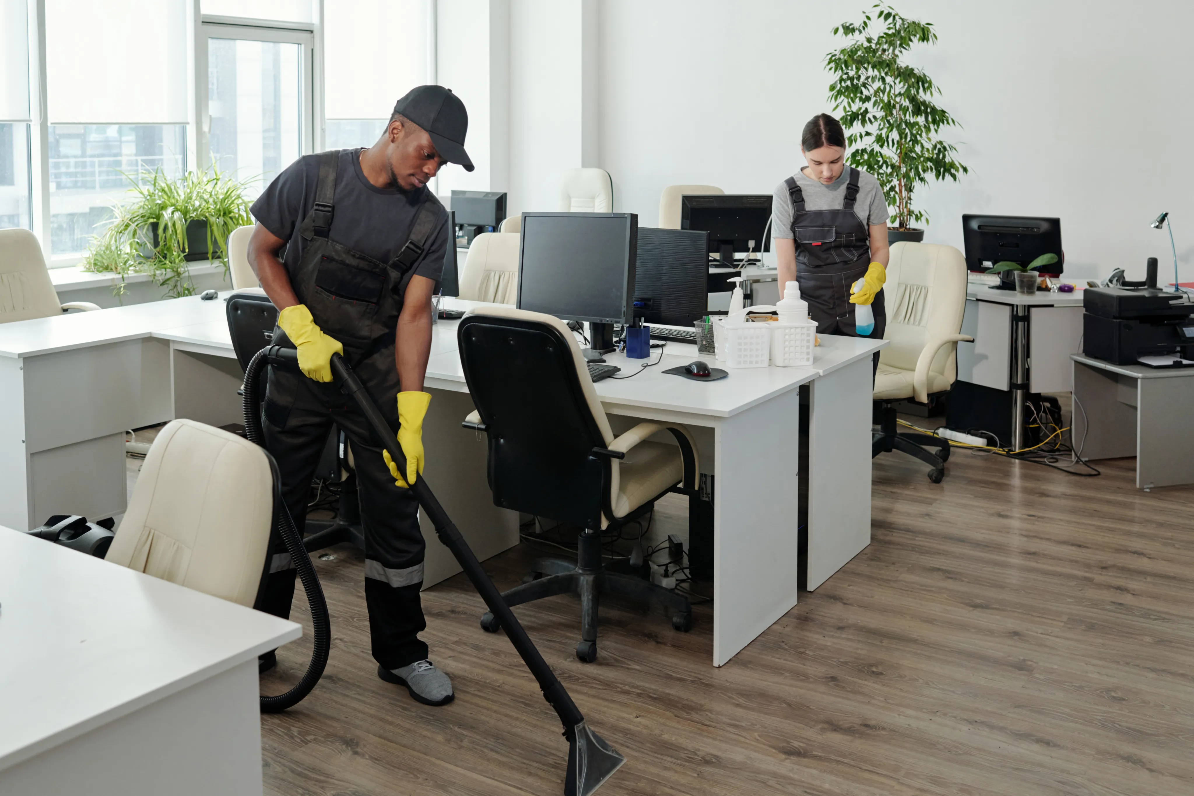 Two commercial cleaners vacuuming and sanitizing desks in a bright, open-plan corporate office environment.