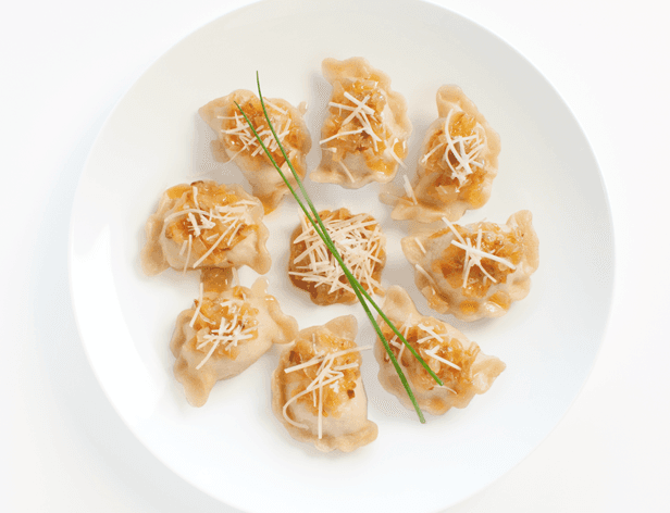 A white plate holds seven dumplings arranged in a circular pattern. Each dumpling is topped with a sprinkling of white shredded cheese and a sprig of green chives. The dumplings have a pale, translucent appearance with a slightly browned edge. The plate is set against a plain white background.