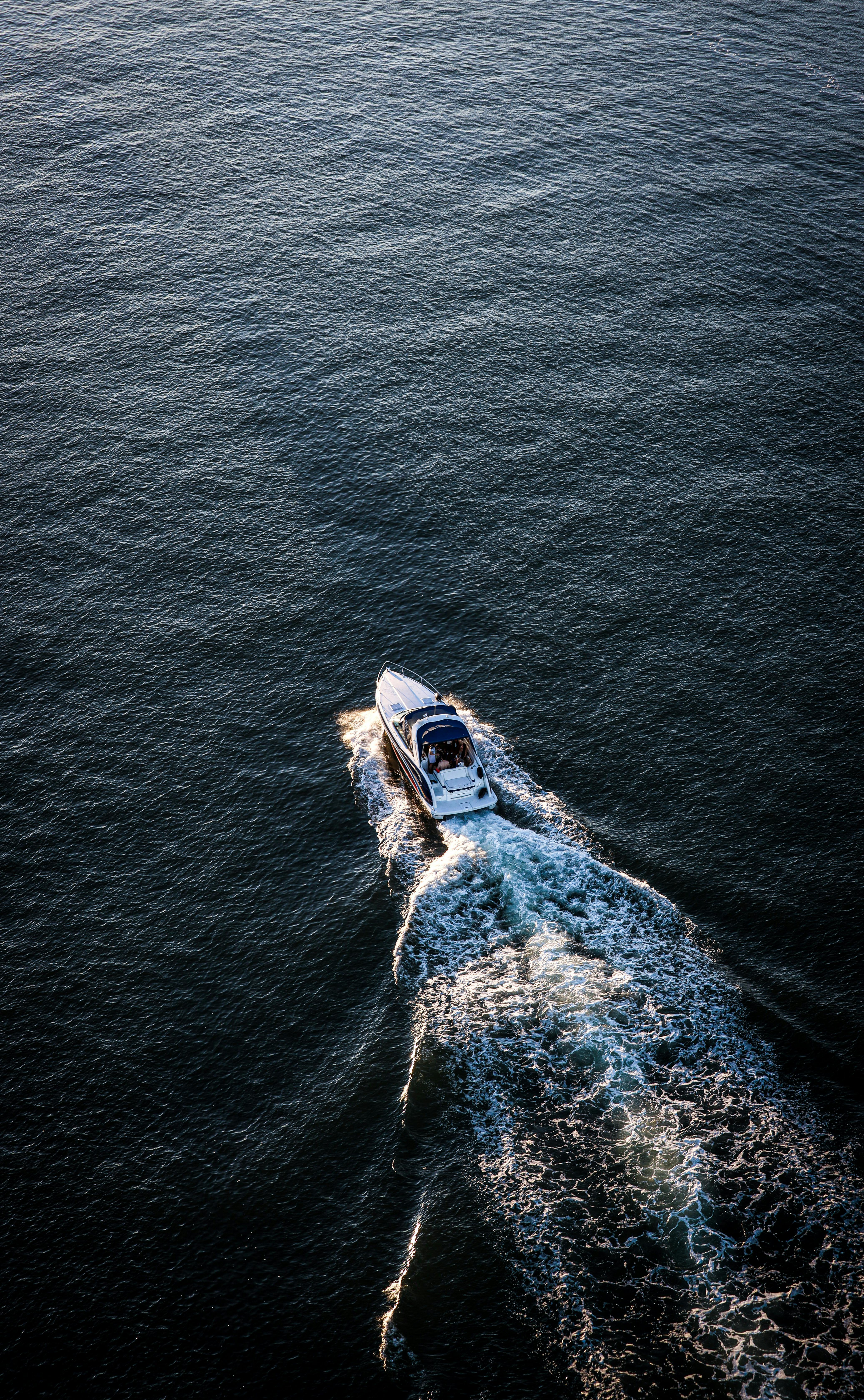 Aerial view of a white speedboat cutting through dark blue ocean waters, leaving a foamy wake behind under a clear sky.