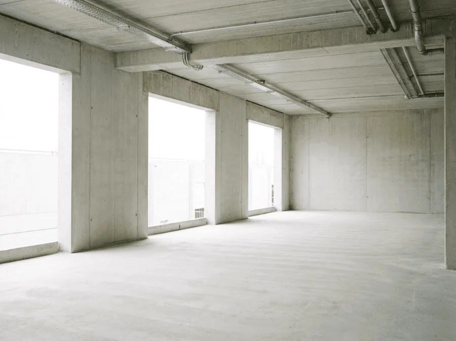 Ein großer, leerer Raum im Rohbauzustand, dessen Wände, Decke und Boden komplett aus rohem, hellgrauem Sichtbeton bestehen. An der linken Wandseite befinden sich drei große, rahmenlose Fensteröffnungen, durch die helles Tageslicht flutet. An der Decke sind freiliegende Installationen wie Kabelpritschen und metallene Rohrleitungen montiert.
