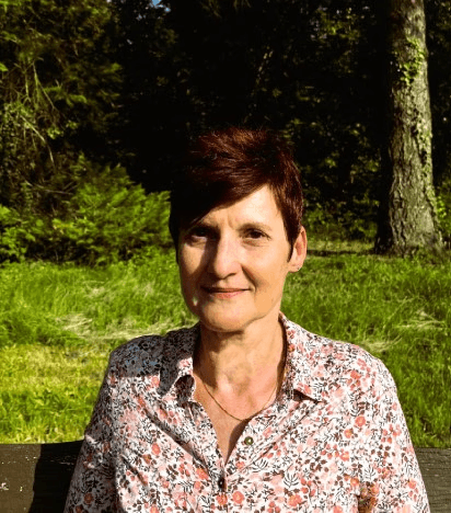 Smiling female therapist with short gray hair, wearing a brown top and a purple shawl, surrounded by a cozy background.