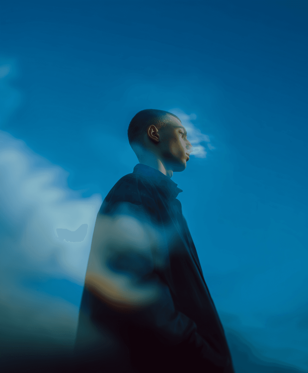 A man with a shaved head looking away from the camera. The image has a blue overcast and clouds around his form.