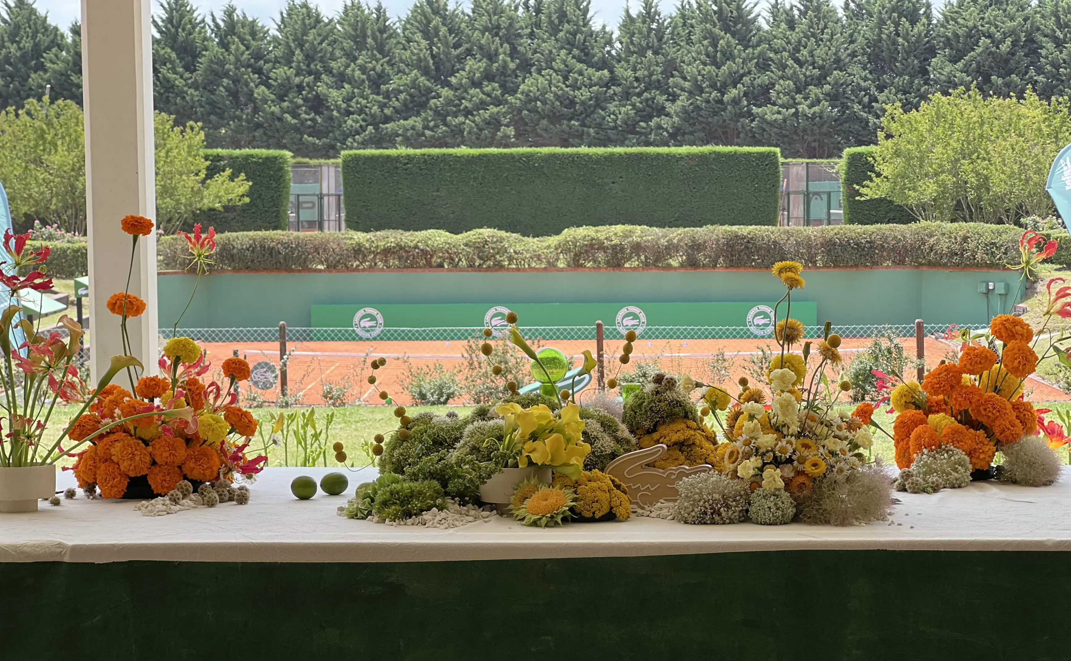 Mesa decorada con arreglos florales en tonos amarillos, naranjas y verdes frente a una cancha de tenis al aire libre, con el icónico logo de Lacoste integrado en la composición central.