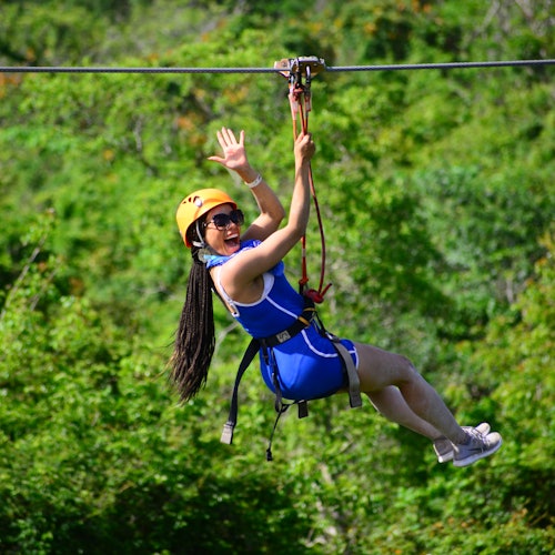 Persona en una tirolesa sobre un exuberante bosque verde, usando casco y arnés, sonriendo y saludando con la mano.