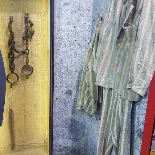 ガラスの後ろに展示されている縞模様の制服と錆びた手錠、コンクリートのような背景が特徴です。