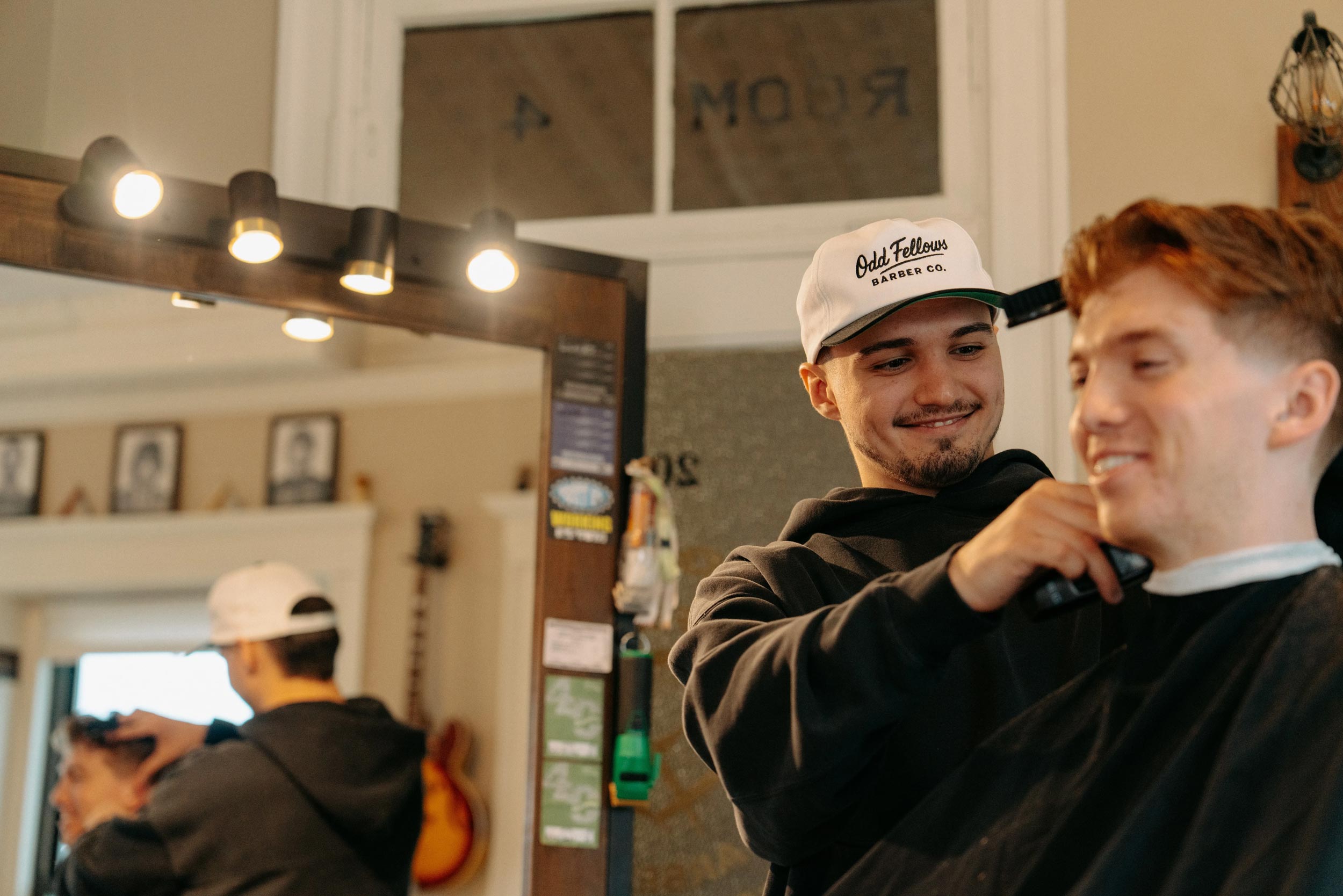 A male barber wearing a white baseball cap with "Odd Fellows Barber Co." on it is cutting the hair of a smiling male client with red hair, who is draped with a black cape. A reflection of the barber working is visible in the mirror behind them.