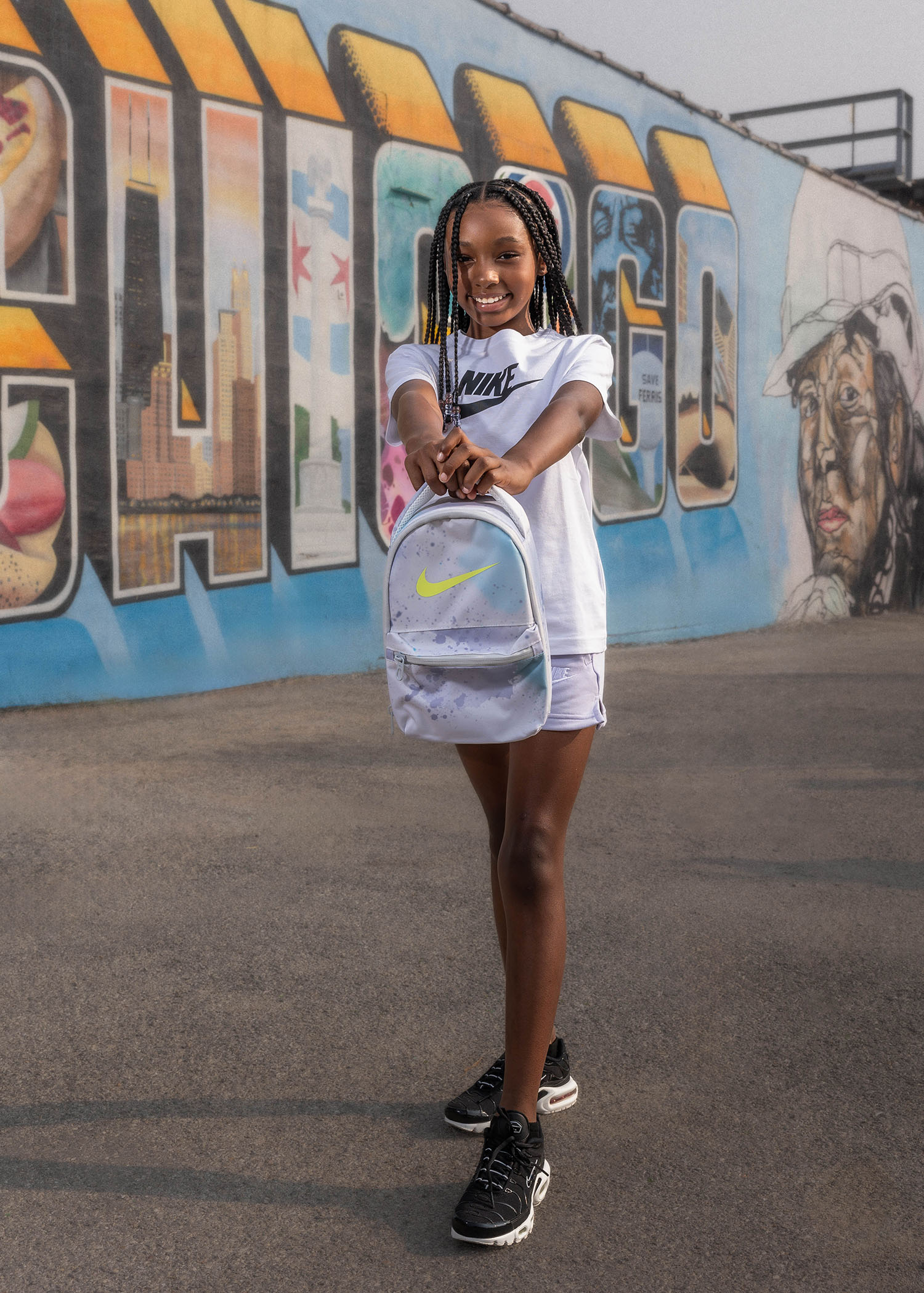 Quickie Studio photographed Nike and Foot Locker’s Chicago community campaign, highlighting youth expression and local culture through vibrant street art and lifestyle imagery.son with braided hair stands smiling and holding a white Nike backpack, standing in front of a vibrant graffiti mural in an urban setting.