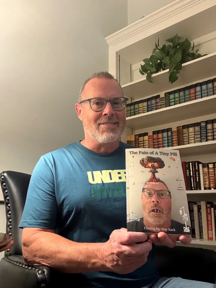Stu holding a copy of his book “The Pain of a Tiny Pill: Finding My Way Back” in front of a bookshelf