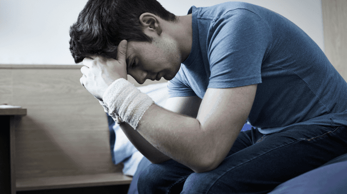 A young man in a blue t-shirt sits hunched over on a bed in a darkened room, head bowed into his bandaged hands, conveying distress or emotional pain.