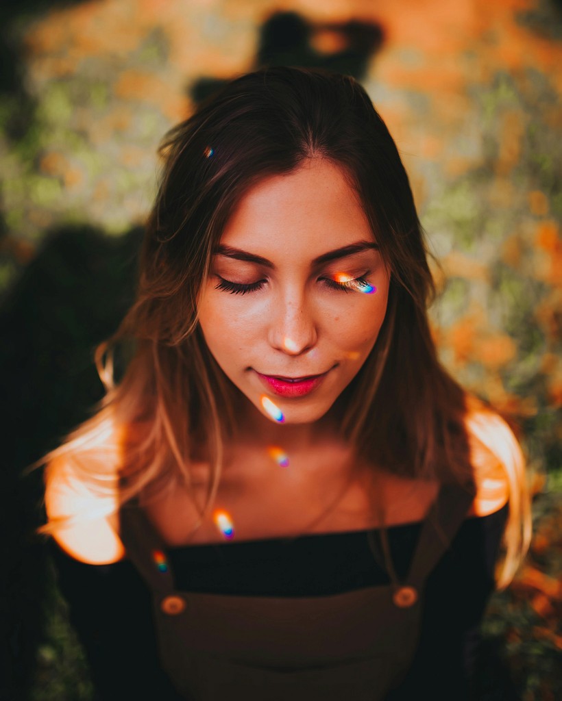 A woman with a white headband relaxing, her face illuminated by warm sunlight filtering through blinds, casting striped shadows. She has a serene and peaceful expression.