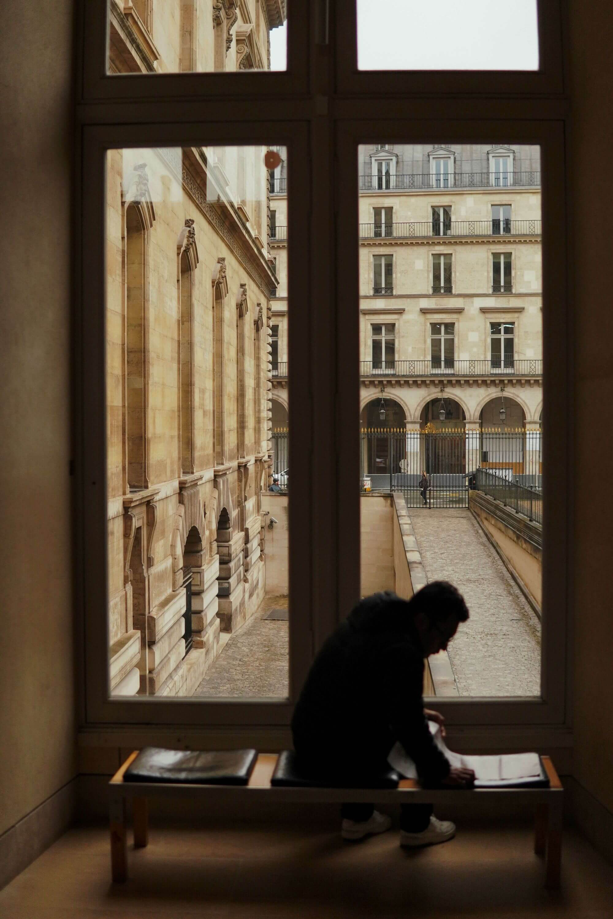 A person sits on a bench inside, silhouetted against a large window. Outside, historic stone buildings with archways and windows are visible, creating a contemplative mood.