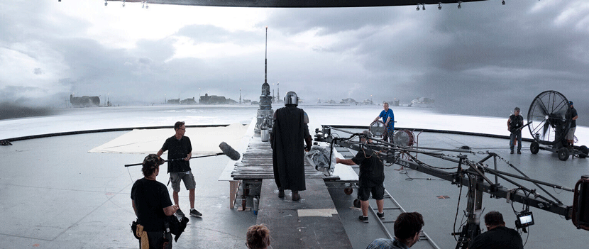 A group of people works on a film set with a cloudy sky in the background, set near a body of water.