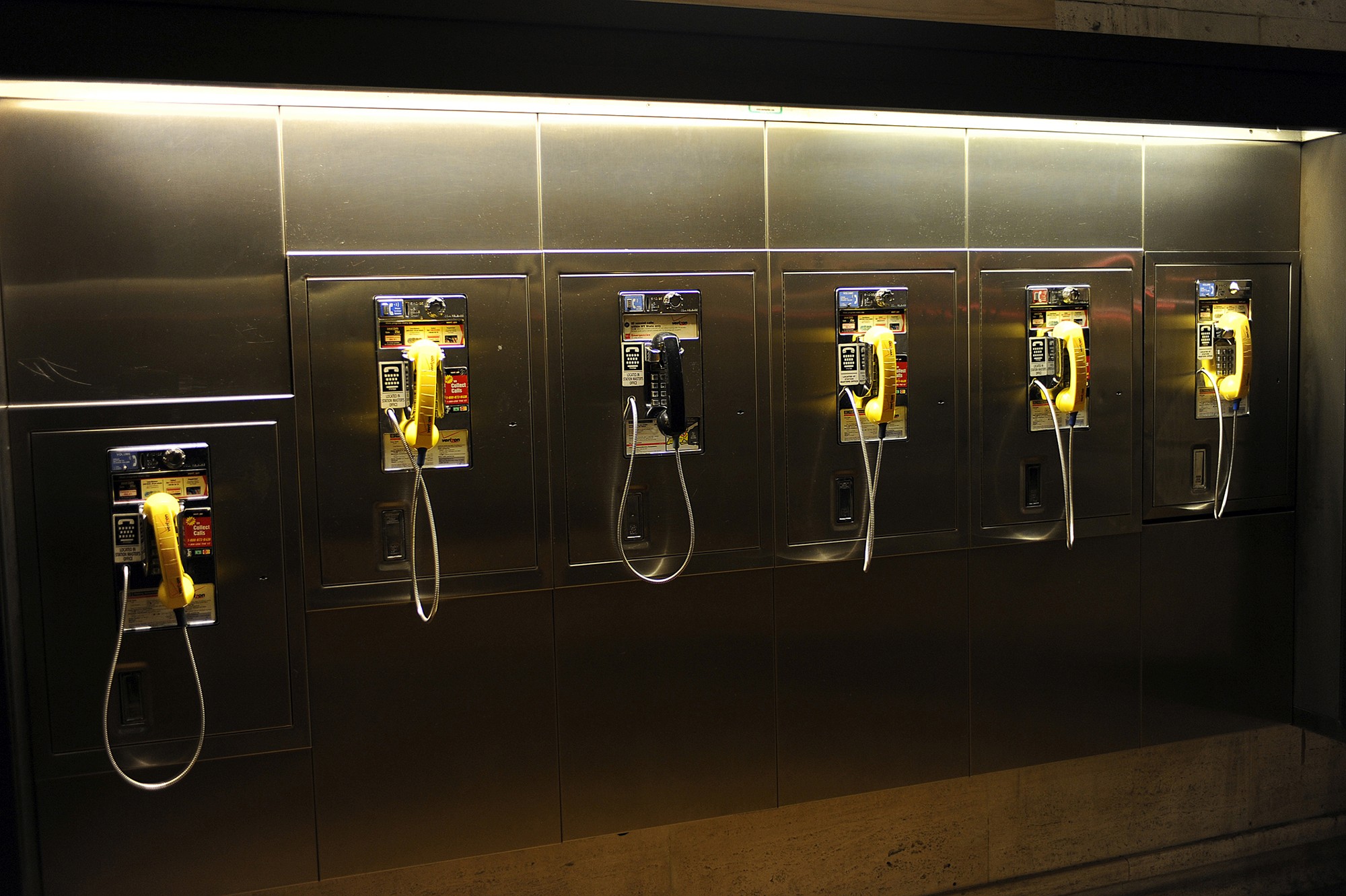 Rangée de cabines téléphoniques murales en métal, photographiées par Frédéric Bourcier dans une lumière chaude et urbaine.