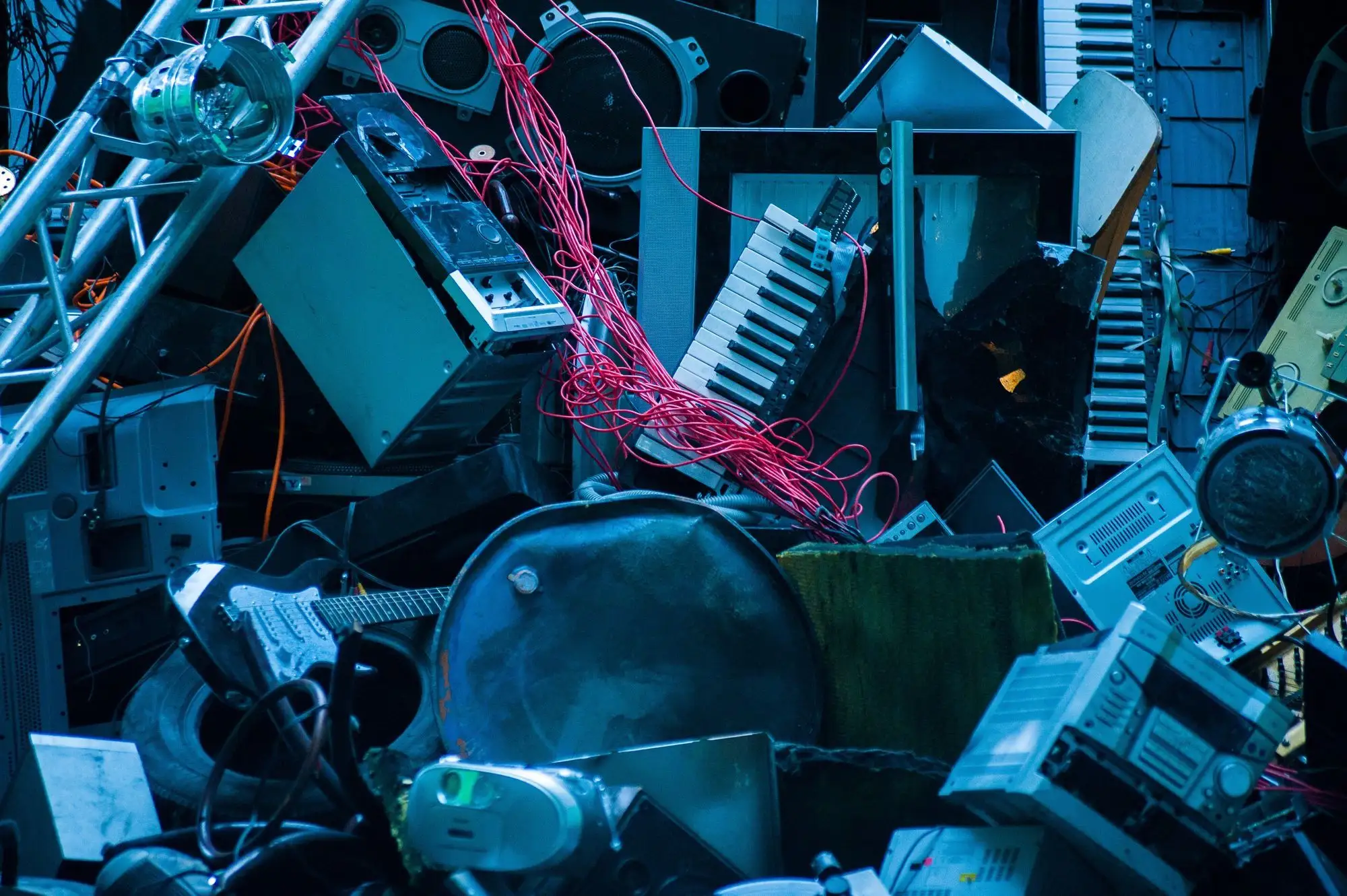 Ein chaotischer Haufen aus alten elektronischen Geräten, darunter Keyboards, Lautsprecher, Kabel, Verstärker und eine E-Gitarre, in blauem Licht beleuchtet – symbolisch für Elektroschrott oder Recycling von Elektronik.