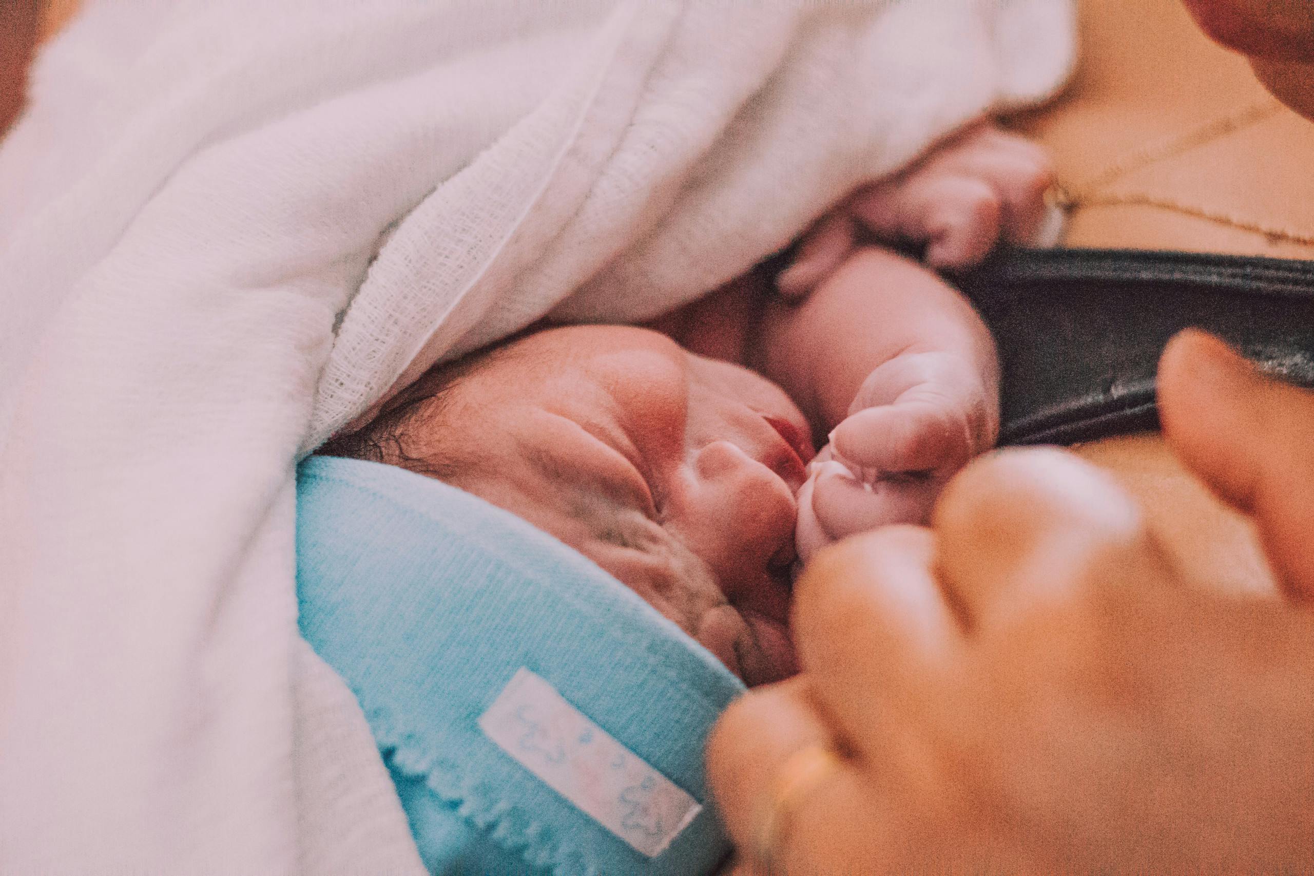 A close up of a newborn's face while it's swaddled in cloth against its mothers chest. Its mother is touching it with her hand. The baby is wearing a light blue hat.