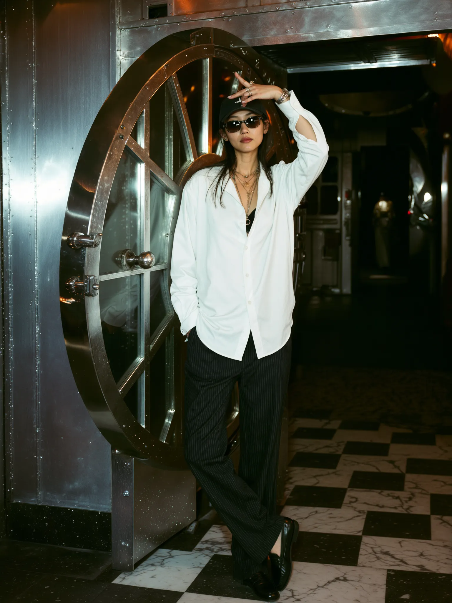 Weibliches KI-Model lässig vor großem Metalltresor, trägt weiße Bluse, dunkle Hose, Sonnenbrille und Accessoires, cooler Nachtclub-Look.