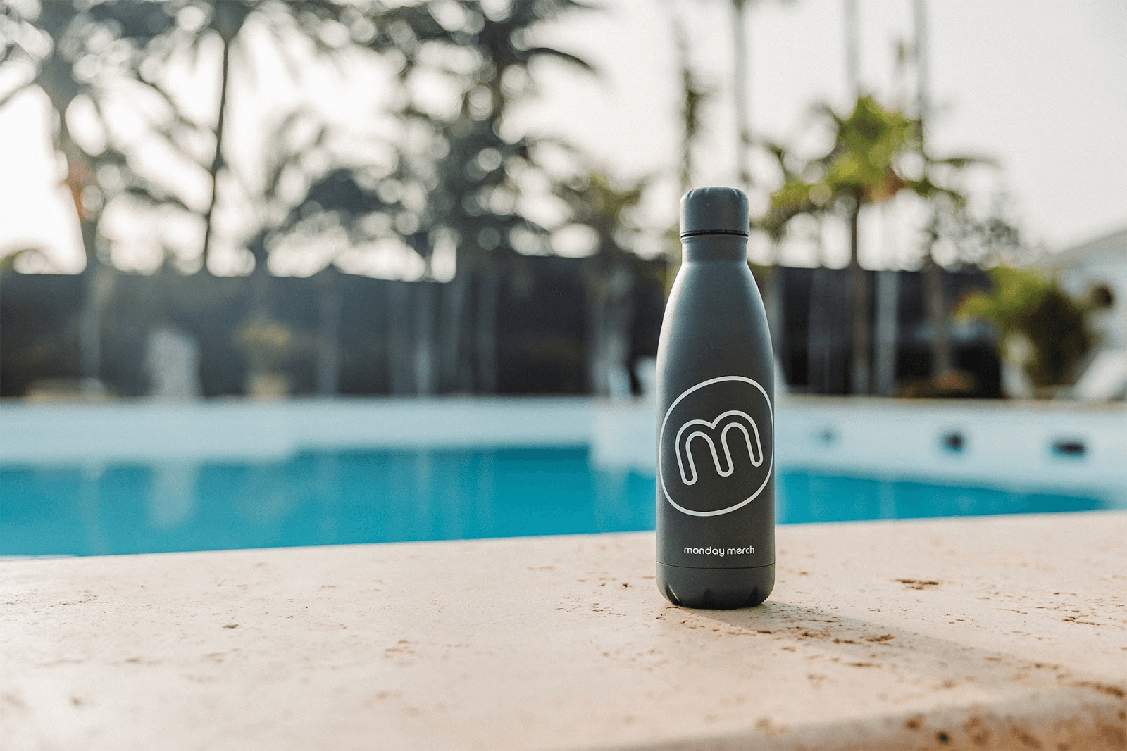 A dark-colored reusable water bottle with a white "m" logo inside a circle and the text "monday merch" below it. The bottle is placed on a ledge in front of a swimming pool, with palm trees and blurred outdoor scenery in the background.