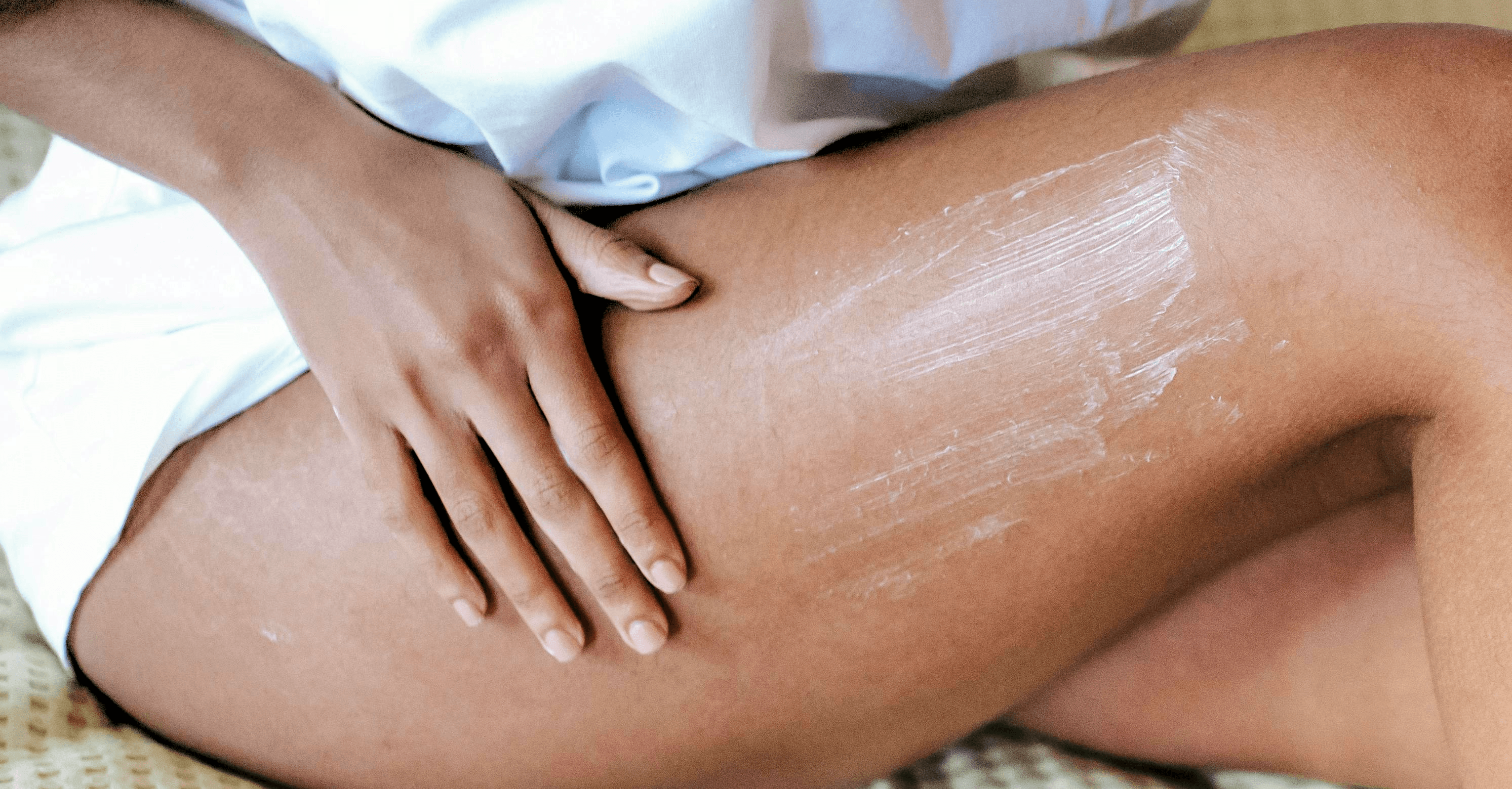 A woman applying lotion to their thigh, wearing a white garment, capturing a moment of skincare and self-care with a focus on the smooth, moisturized skin