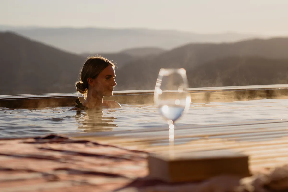 Frau im Infinity-Pool bei Sonnenuntergang, davor ein Weinglas und ein Buch am Beckenrand