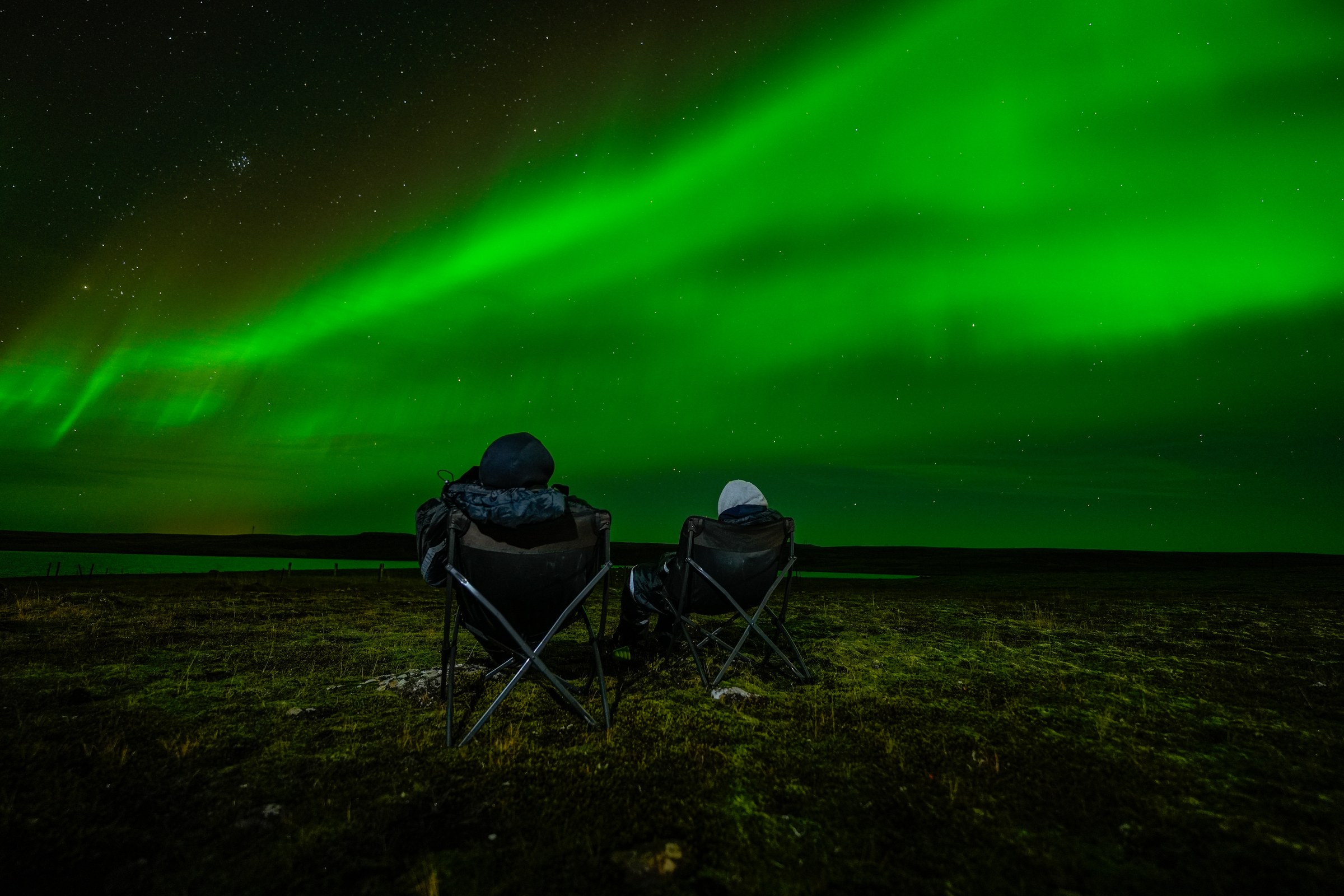 Two guests sitting in camp chairs on open ground watching a green aurora band overhead