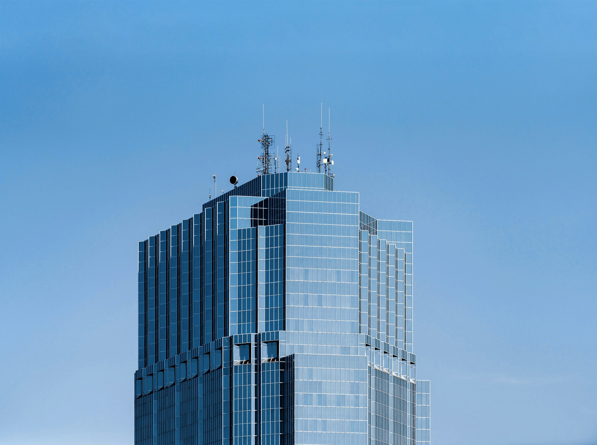 Modern glass building with antennas and structures on the rooftop under a clear blue sky.