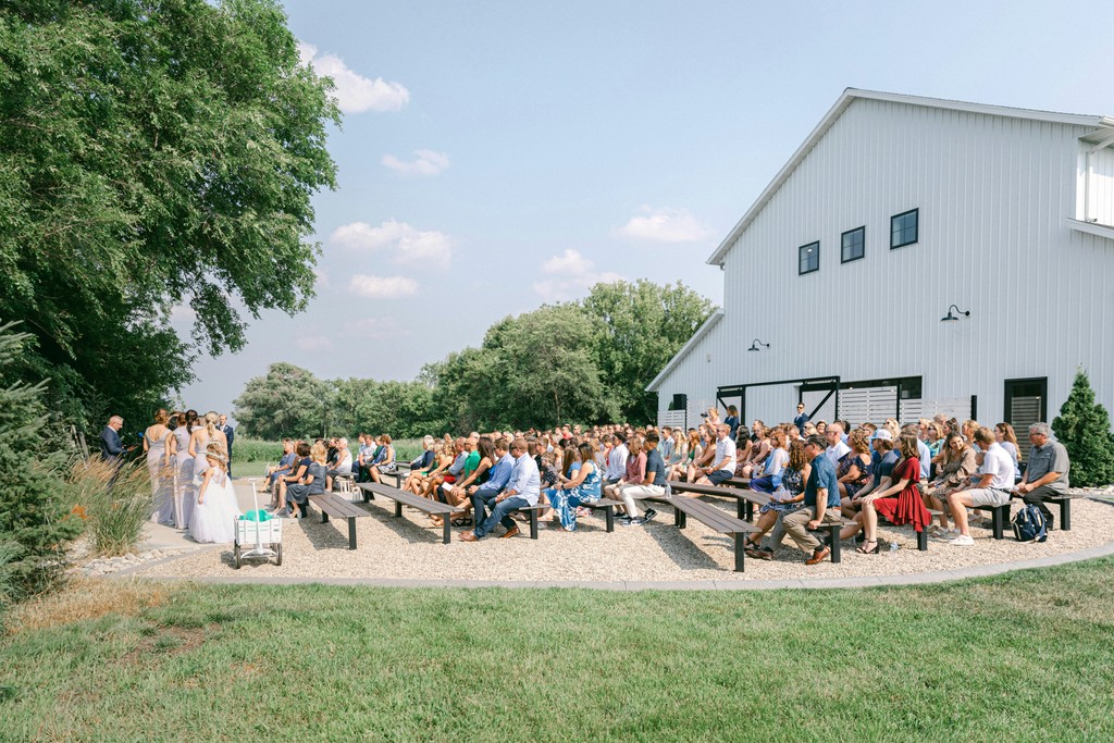 Outdoor wedding ceremony with guests on wooden benches beside a modern white barn venue on a sunny summer day