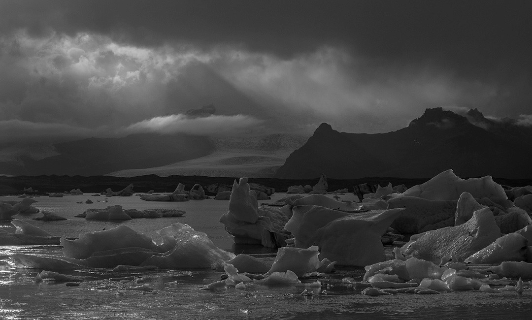 Icebergs floating in a glacial lagoon beneath dark clouds and distant mountains.