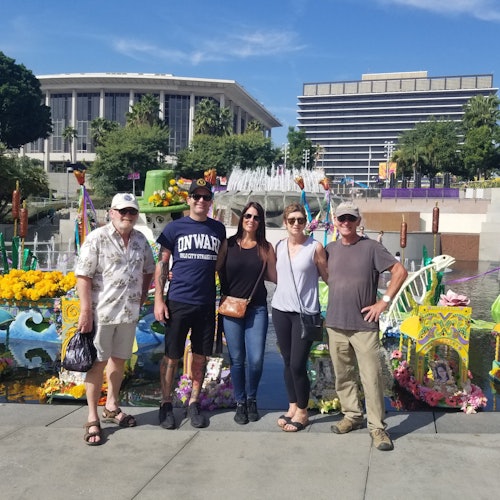 Five people standing in front of colorful flower decorations, with fountains and modern buildings in the background on a sunny day.