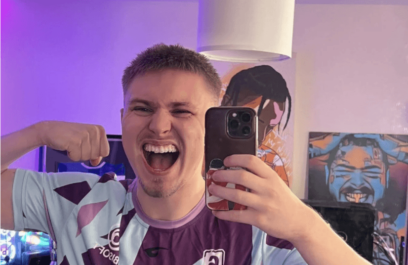 Ein junger Mann macht ein Selfie im Spiegel, trägt ein lila-hellblaues Trikot und posiert lachend mit angespanntem Bizeps. Im Hintergrund sind bunte Wandbilder und ein Computer-Setup zu sehen.