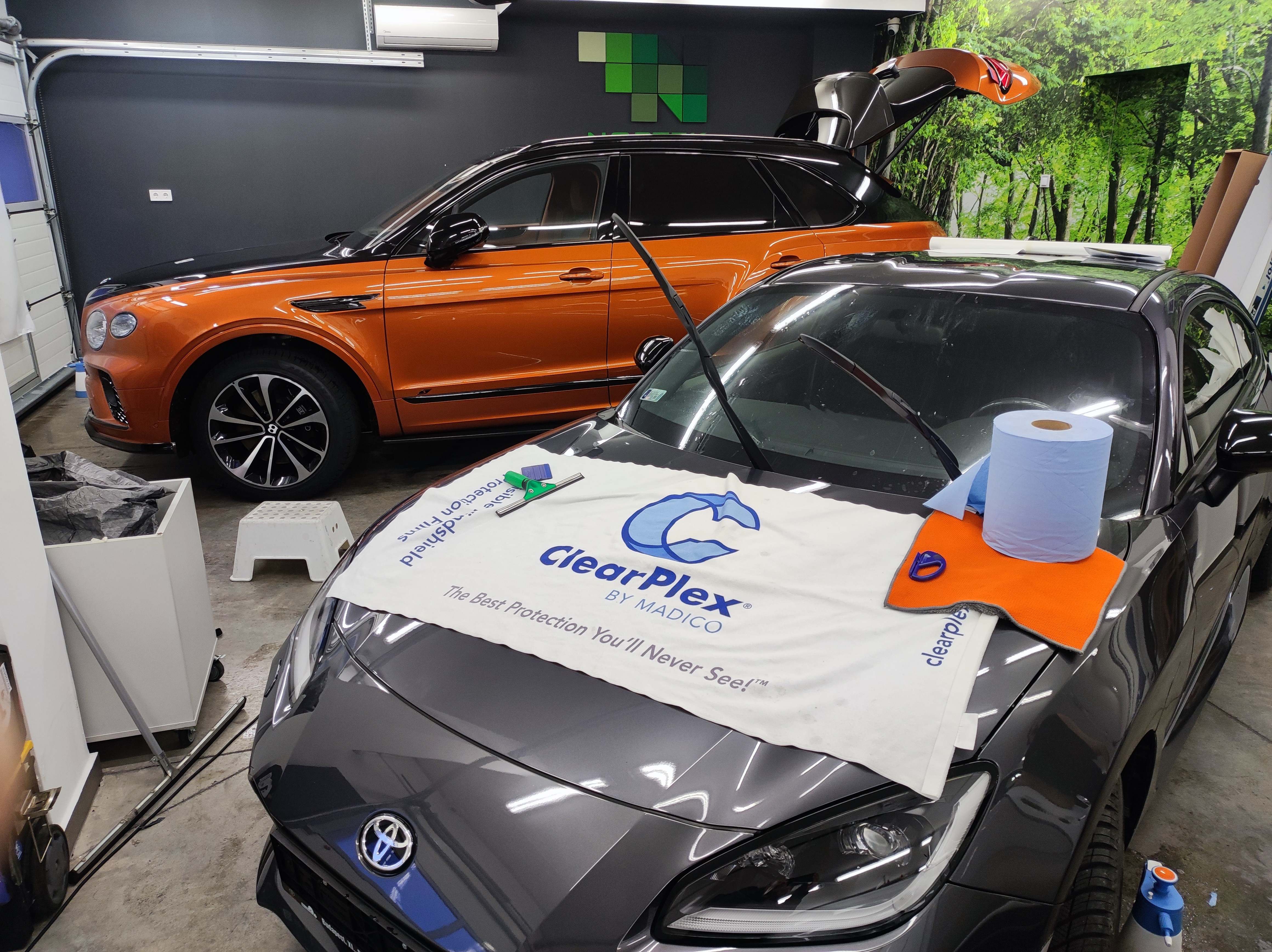 ClearPlex szélvédővédő fólia felhelyezésének előkészületei egy szürke Toyota GT86 sportautón, a háttérben egy kéttónusú Bentley Bentayga luxus terepjáróval a Nortek stúdiójában.