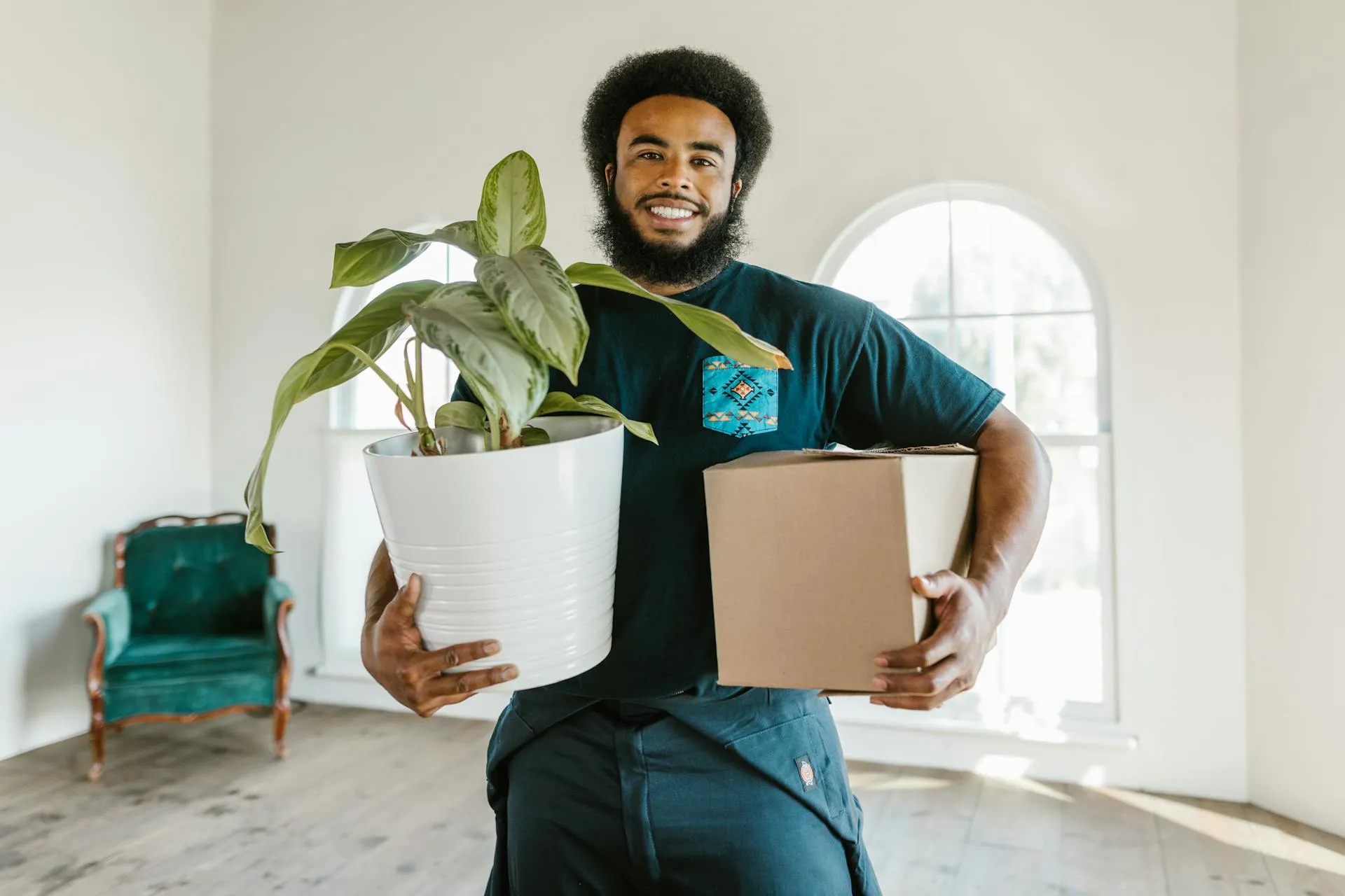 Smiling man holding a potted green plant and a cardboard box inside a bright room during a home relocation.