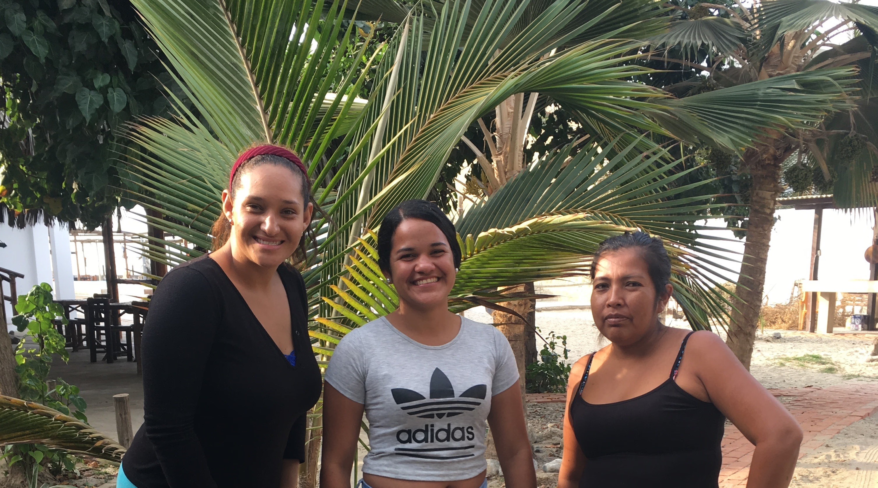 Three women smiling together in front of tropical palm trees.
